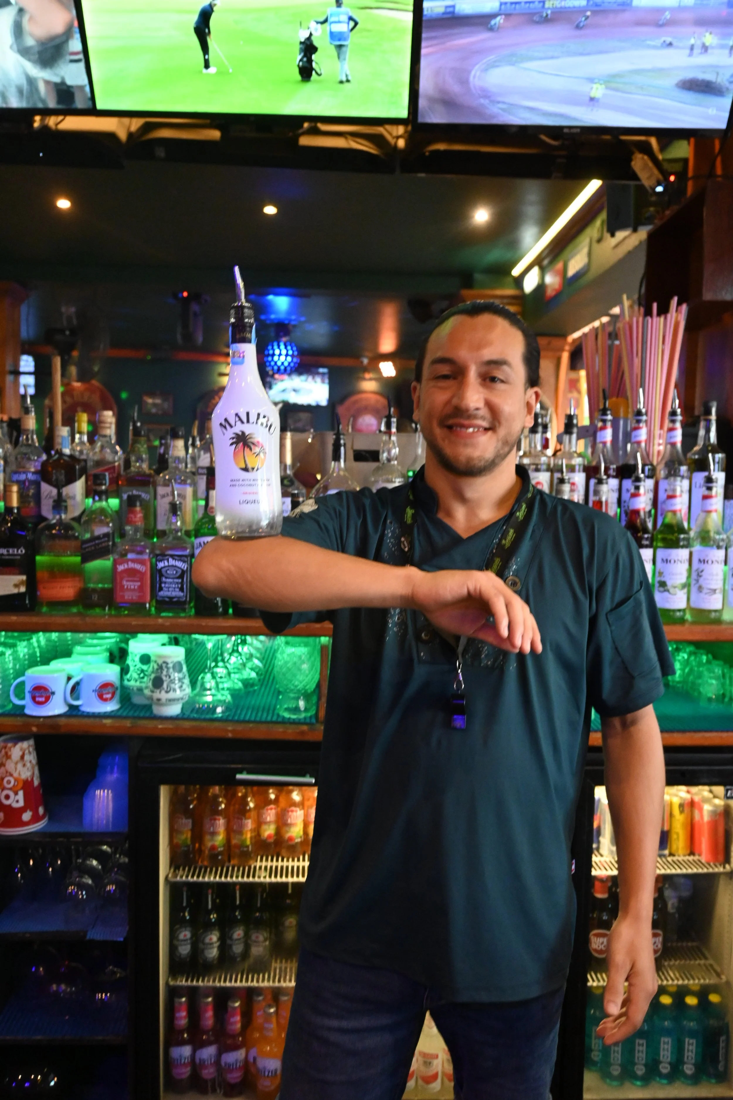 A bartender standing behind a bar, holding a Malbec wine bottle on his arm, smiling. The bar is stocked with various bottles of liquor and colorful cups, with a TV mounted on the wall showing sports.