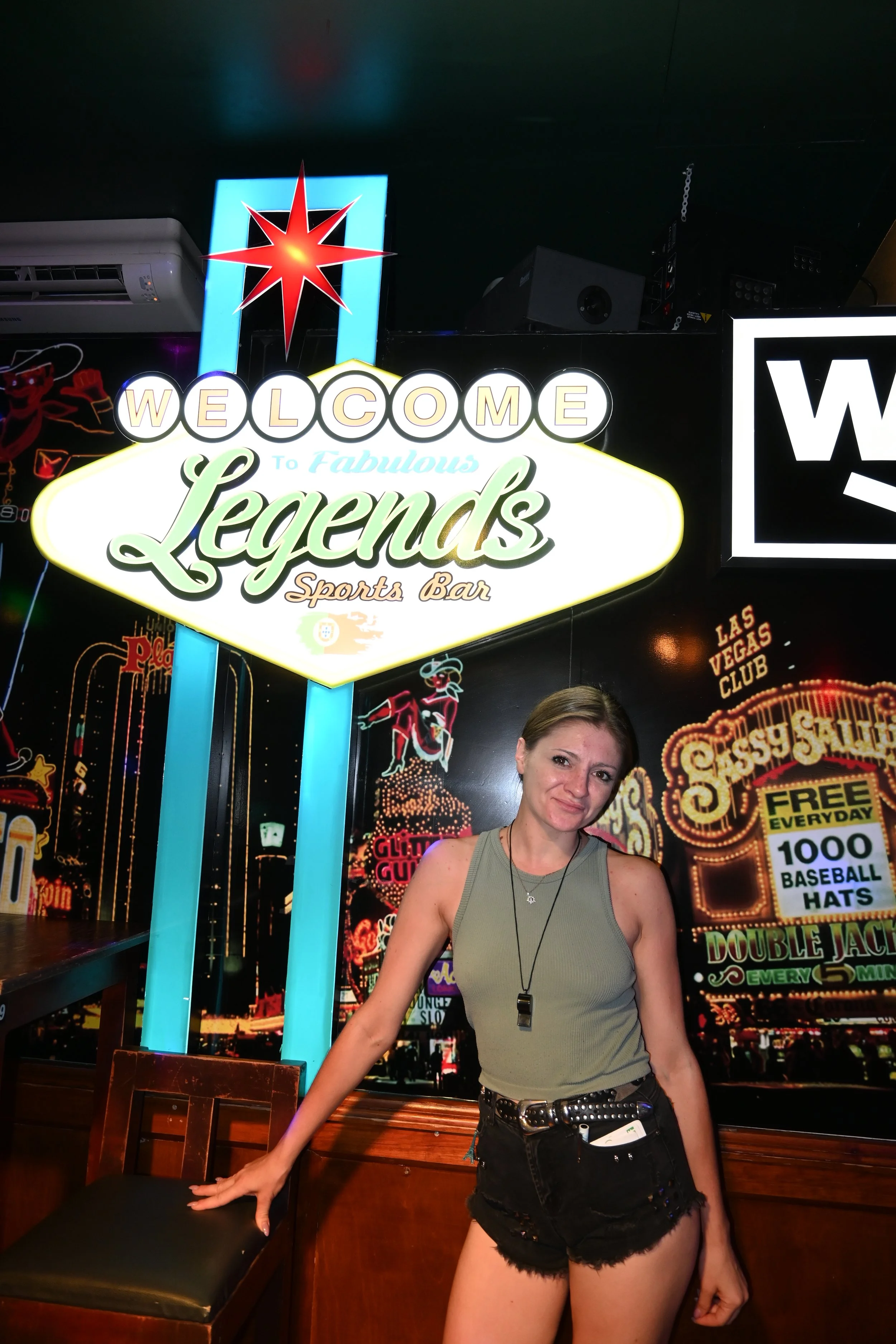 A woman standing in a lively bar or club, with colorful neon signs and bright lights in the background. She is wearing a sleeveless top, black shorts, and a headset or necklace, with her right hand resting on a chair.