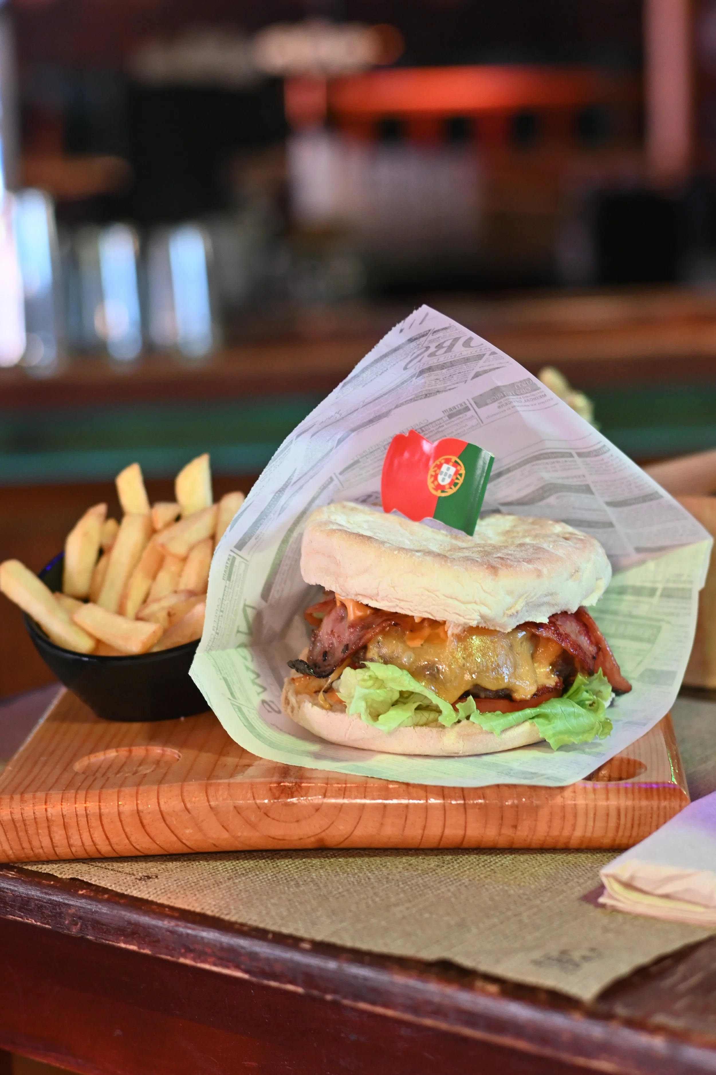 A sandwich with bacon, cheese, lettuce, pickles, and sauce on a bun, served with a side of French fries on a wooden tray.