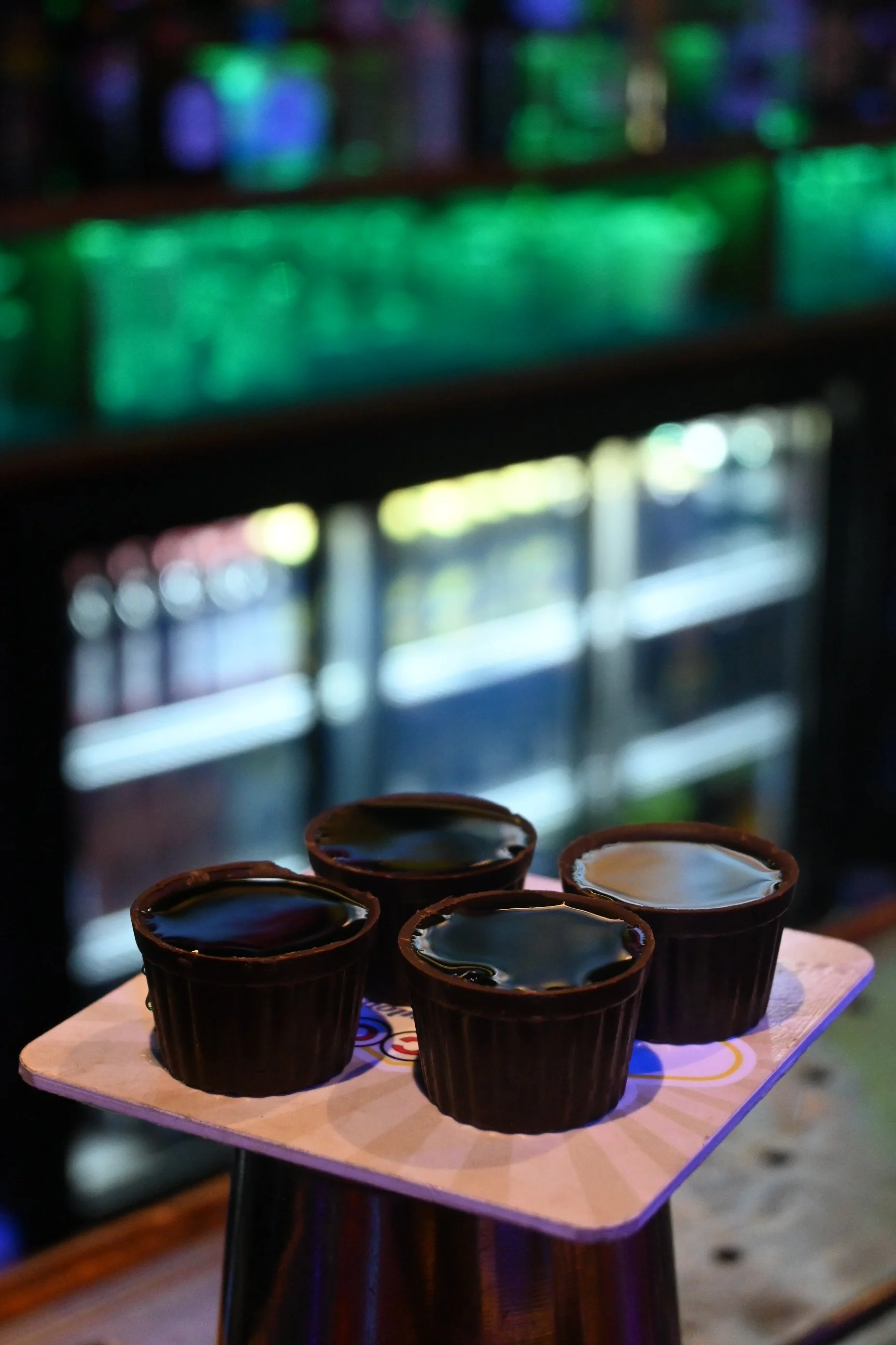 Four small chocolate cups filled with dark liquid, placed on a square white tray with colorful patterns, in a dimly lit environment with blurry bottles and shelves in the background.