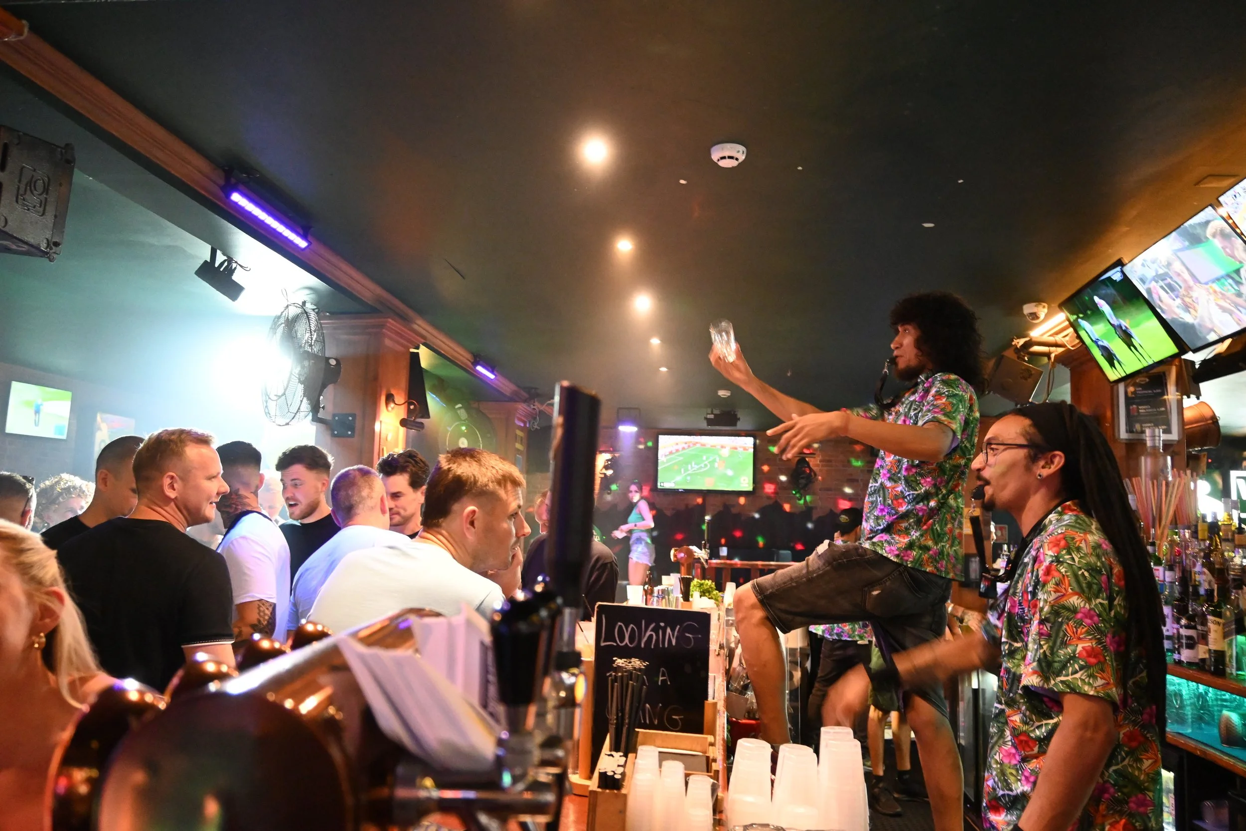 People enjoying a lively night at a bar with a live performer on stage, wearing colorful Hawaiian shirts, with multiple television screens showing sports in the background.