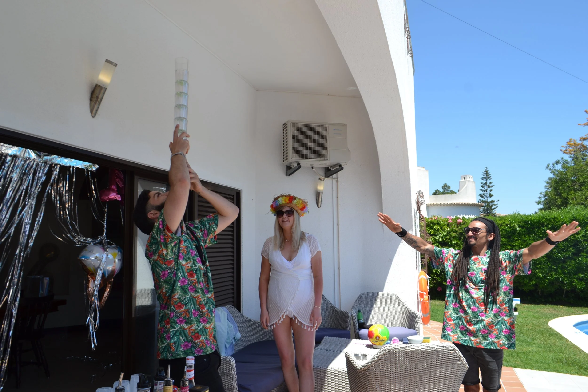 People celebrating outdoors, some wearing colorful Hawaiian shirts and a woman in a white dress, with party decorations and balloons on a sunny day.
