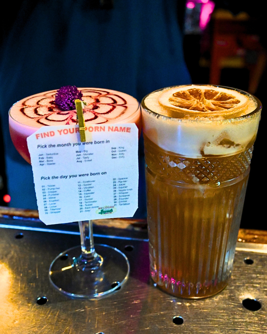 Two cocktails on a bar counter: one pink drink with a purple flower garnish and a find your zodiac sign card, and the other is a lemon drink with a lemon slice on top.
