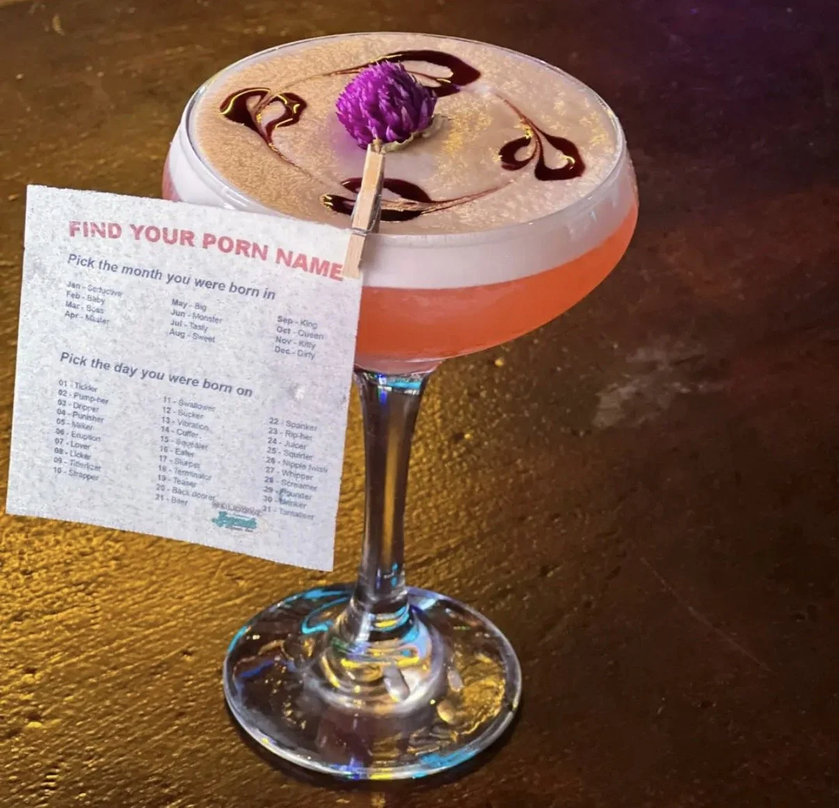 A pink cocktail garnished with a purple flower and chocolate drizzle, with a paper clip attached to a printed sheet titled 'Find Your Porn Name' hanging from the glass. The sheet contains fun prompts about birth month and date.