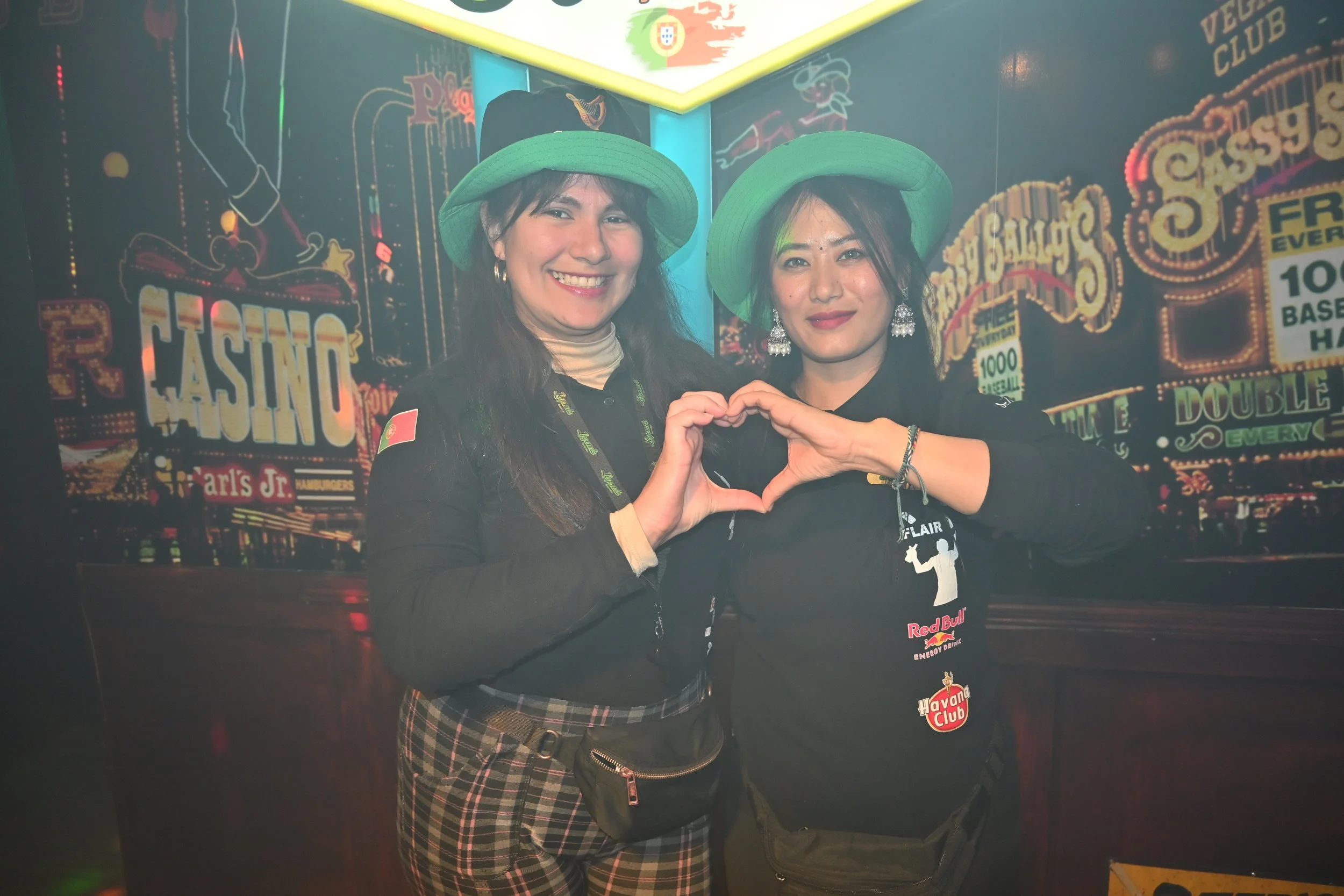 Two women wearing green hats are standing together, making a heart shape with their hands. They are smiling in front of a themed background with neon signs and phrases like 'Casino' and 'Havana Club'.