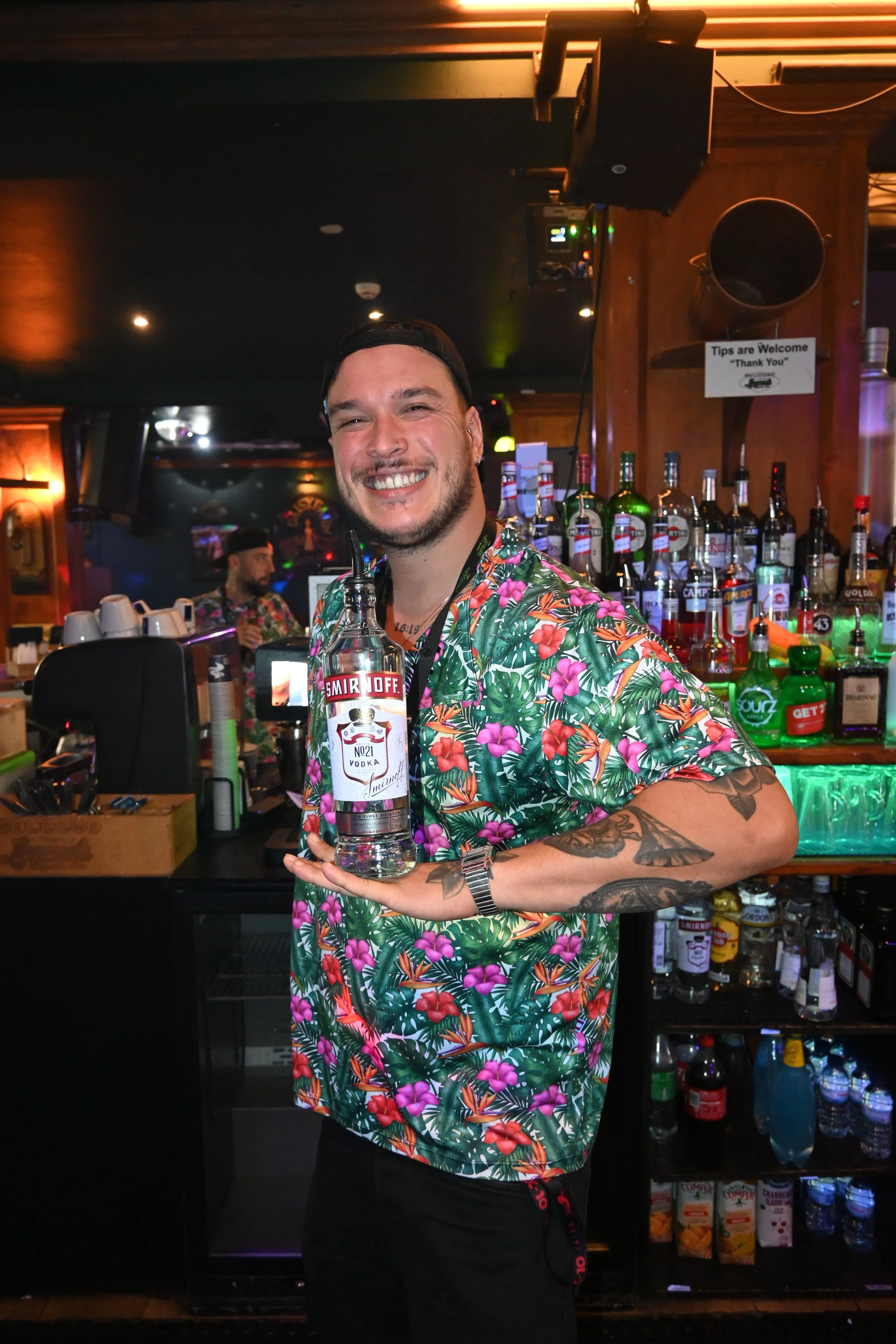 A smiling man with tattoos wearing a floral shirt holding a Smirnoff vodka bottle in a bar.