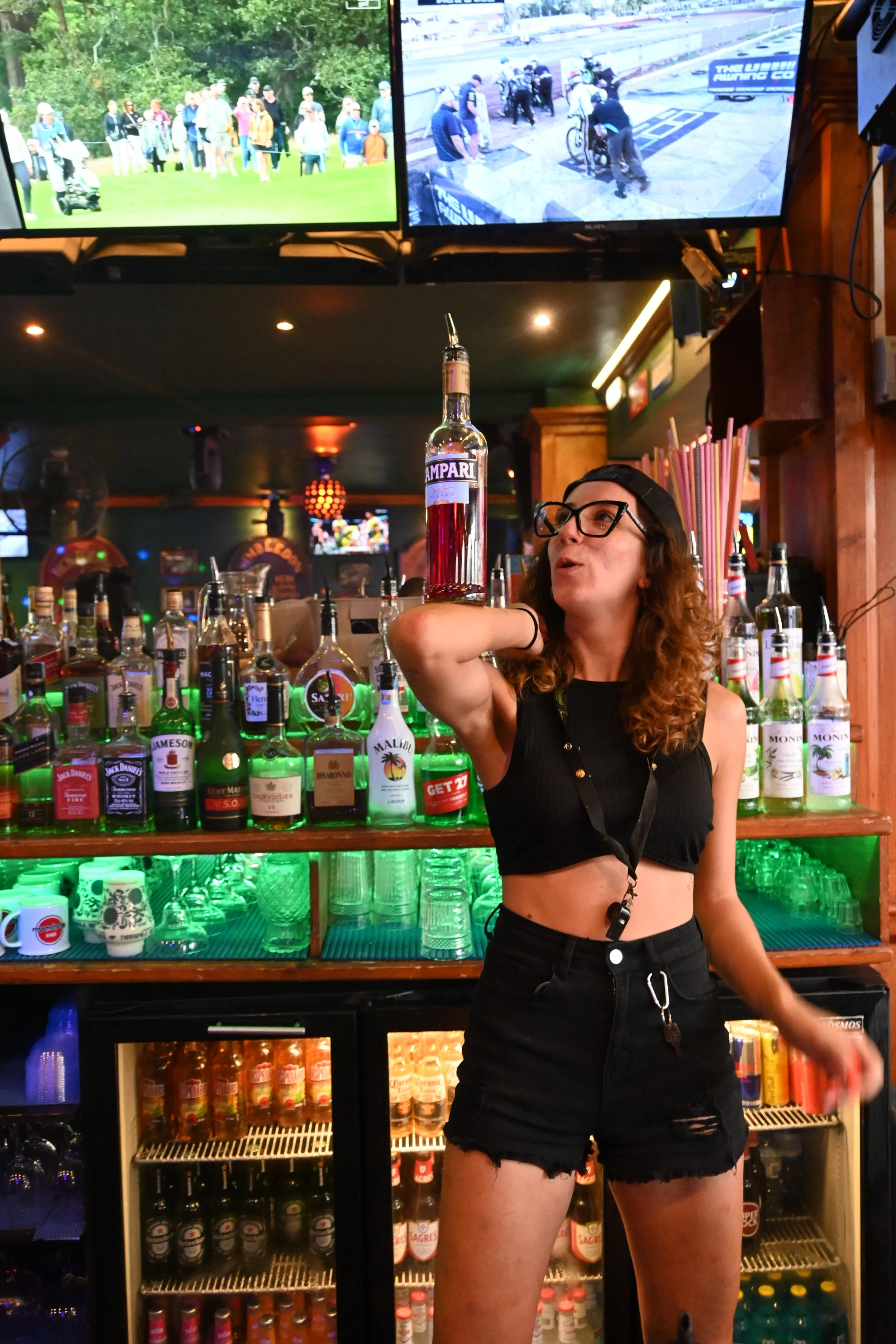 A woman with curly hair, black glasses, wearing a black crop top and black shorts, balancing a large bottle of Campari on her shoulder inside a bar.