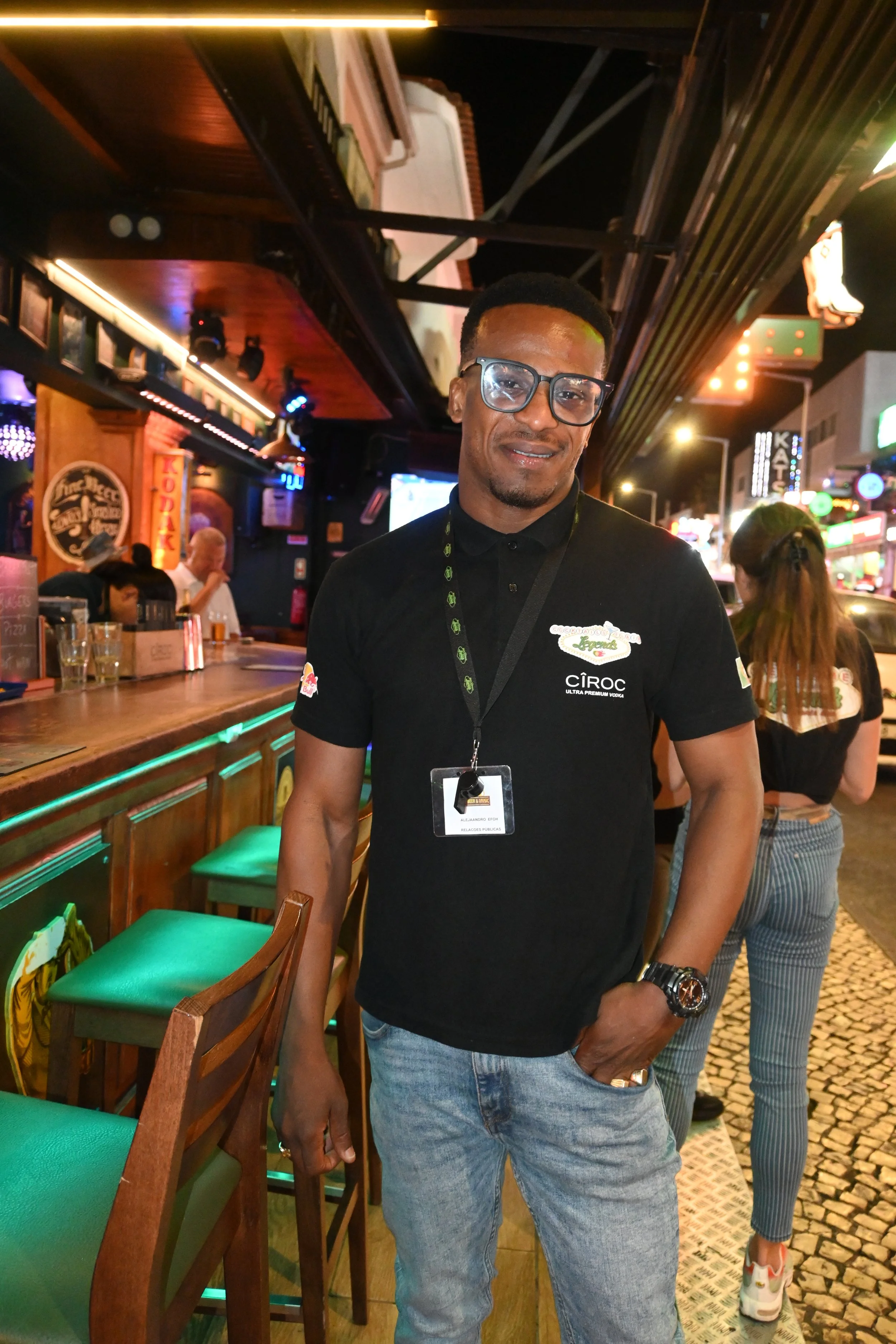 A man wearing glasses, a black polo shirt with logos, and jeans stands at a bar in a nightlife setting. The bar has green cushioned seating and a wooden counter. In the background, other people are socializing, and neon signs and bright lights decorate the night outside.