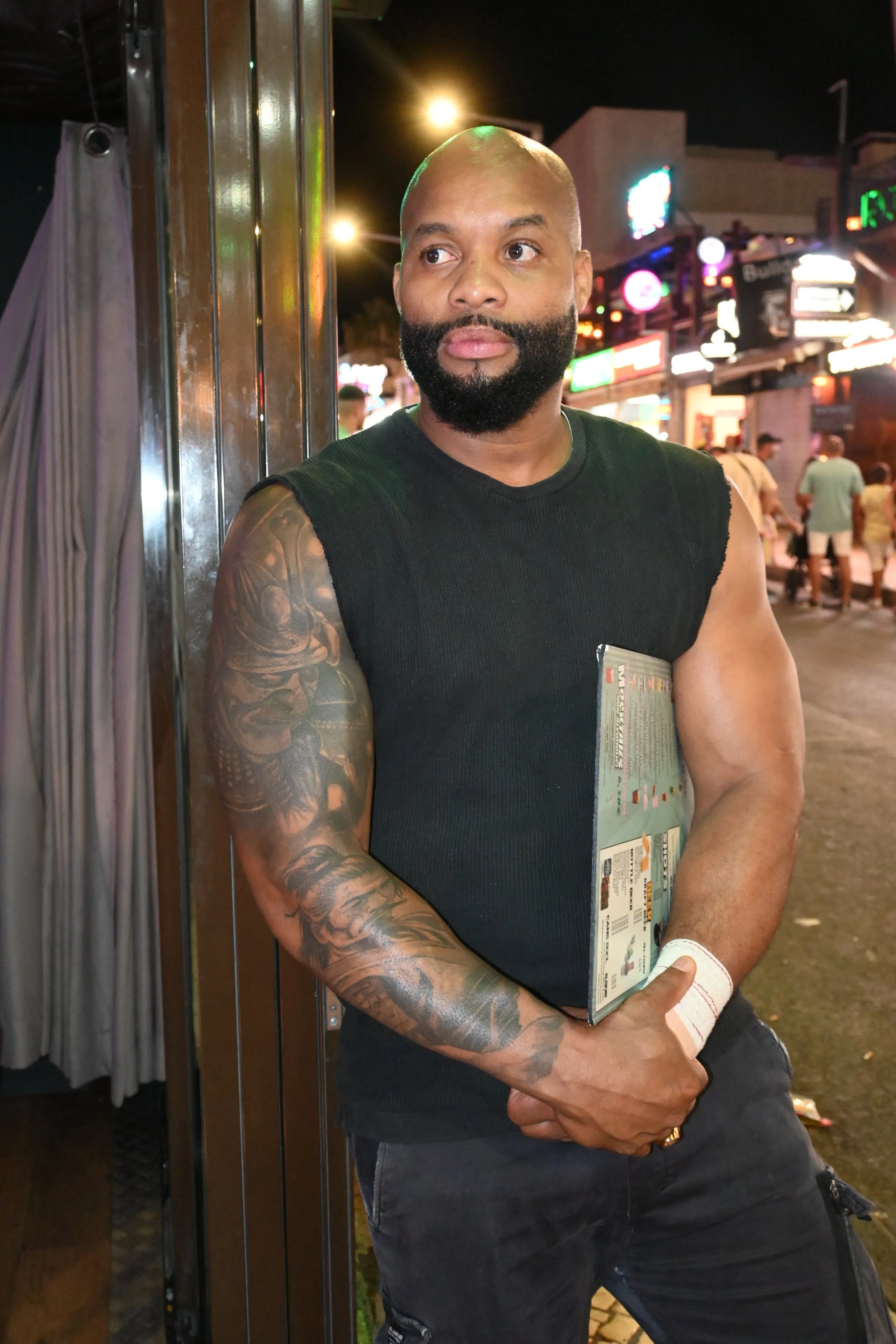 A man with a tattooed arm holding a booklet standing outside at night with a busy street and neon signs in the background.