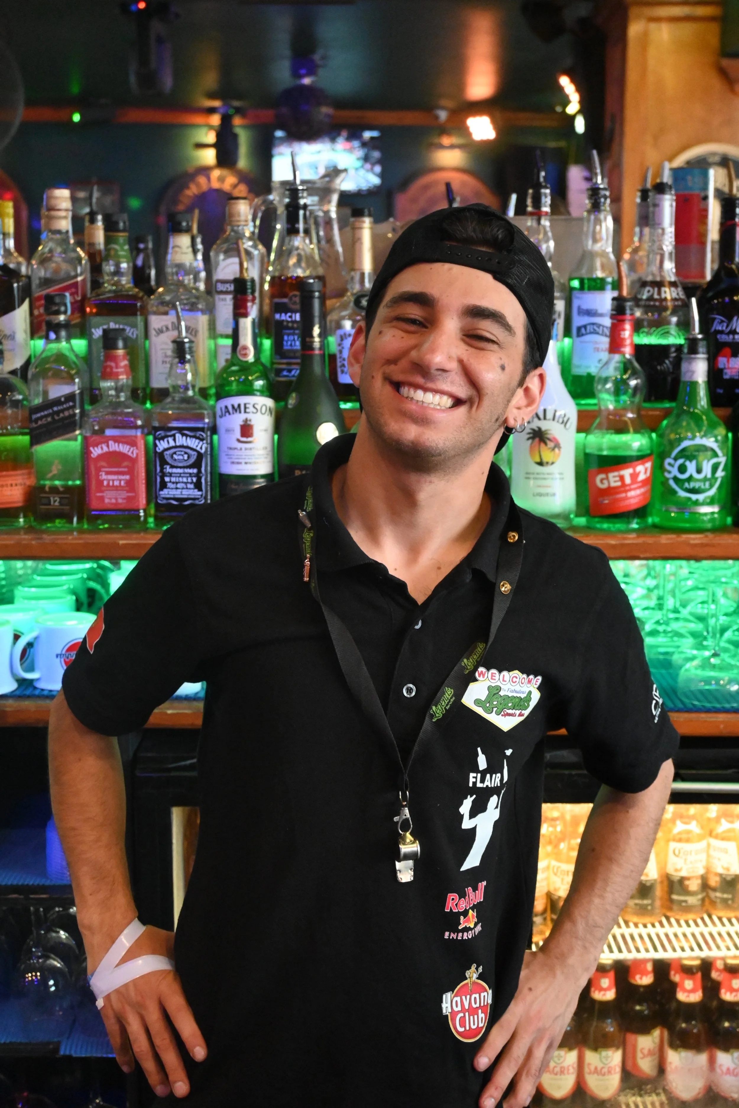 A young man smiling at a bar with colorful liquor bottles behind him.