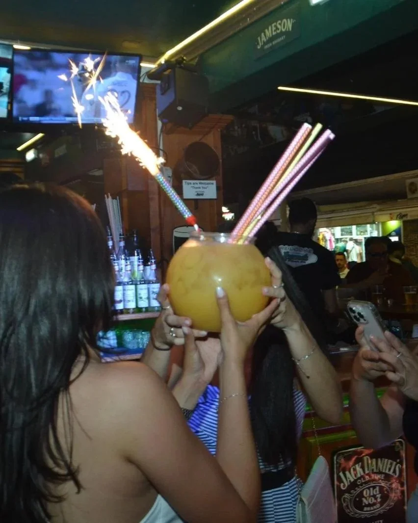 Person holding a yellow coconut with colorful straws inside, and sparklers lit on a sparkler stick sticking out of the coconut, inside a bar or pub with people in the background.