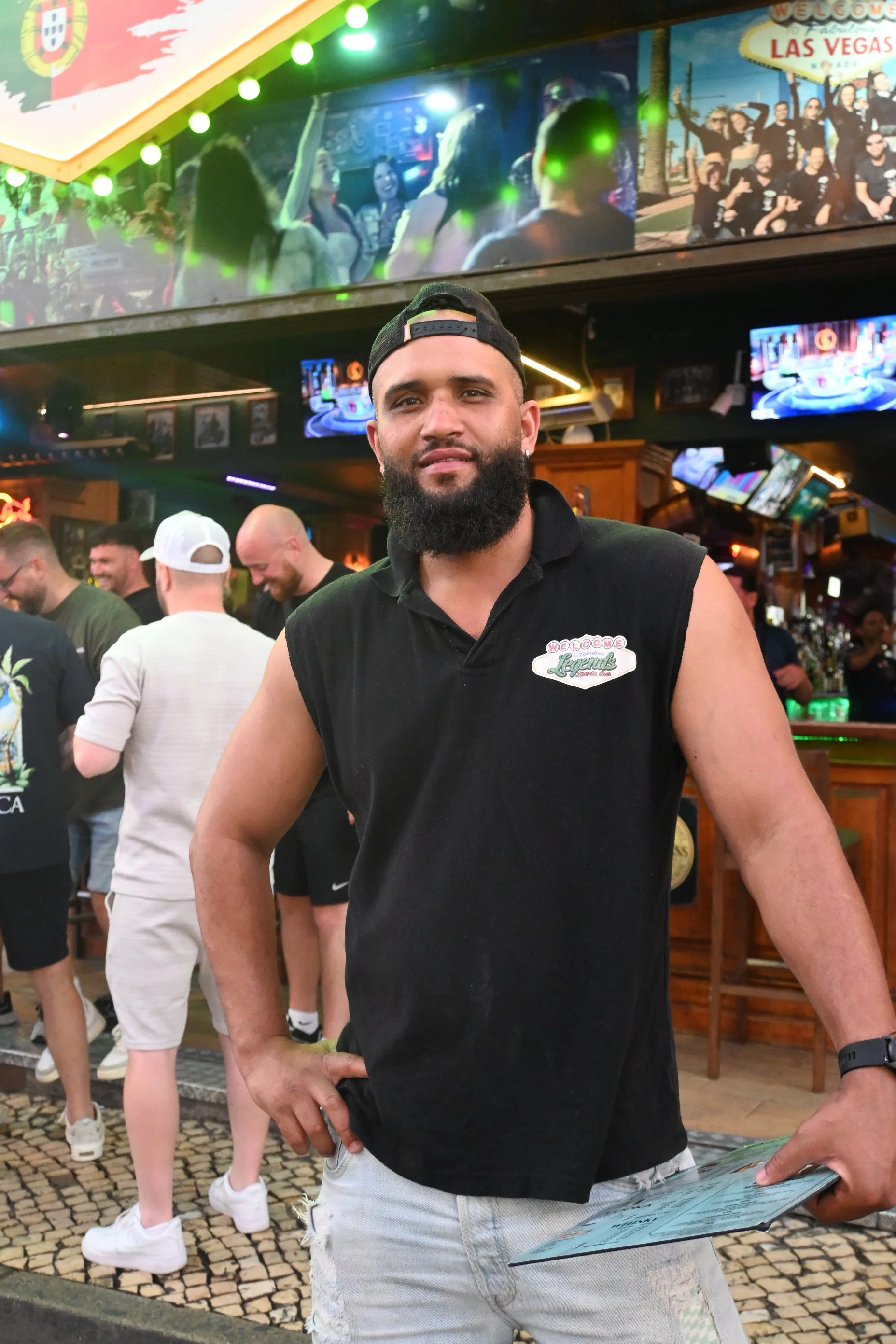 A man with a beard, wearing a black sleeveless shirt with a pin that says "Welcome to Legendary" and a black cap backwards, standing in a lively bar or restaurant with several people in the background. There are multiple televisions and a sign saying "Las Vegas."