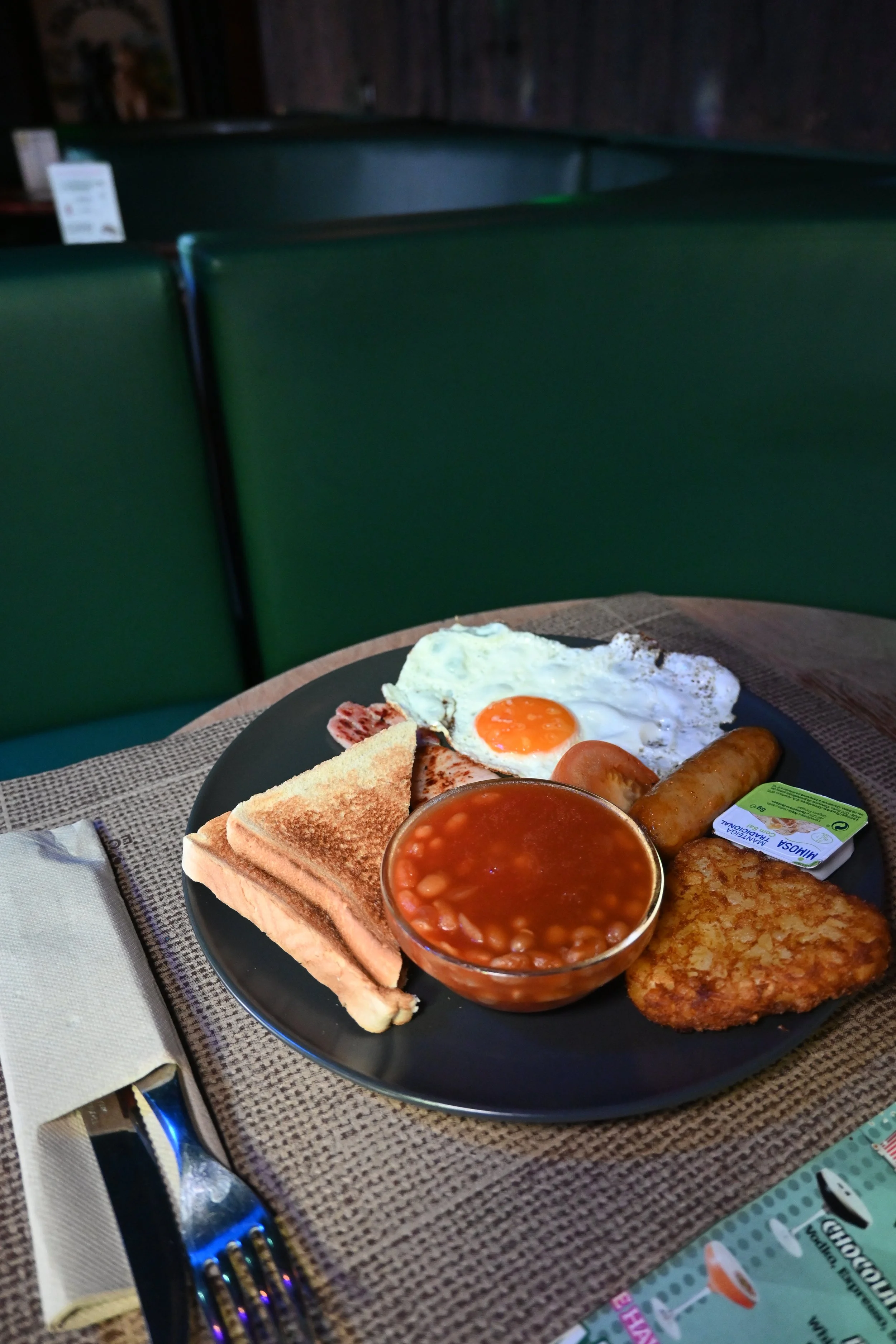Breakfast plate with fried egg, baked beans, toast, sausage, hash browns, and butter on a black plate, on a table with a placemat, fork, and knife.