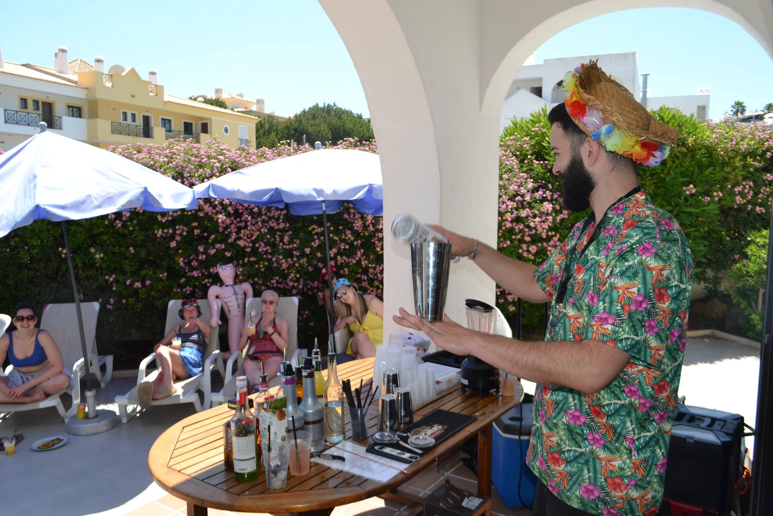 A man with a beard wearing a straw hat decorated with colorful flowers and a tropical shirt, preparing drinks at an outdoor tropical-themed party. Seated guests, umbrellas, and a pink flowering bush are visible in the background.