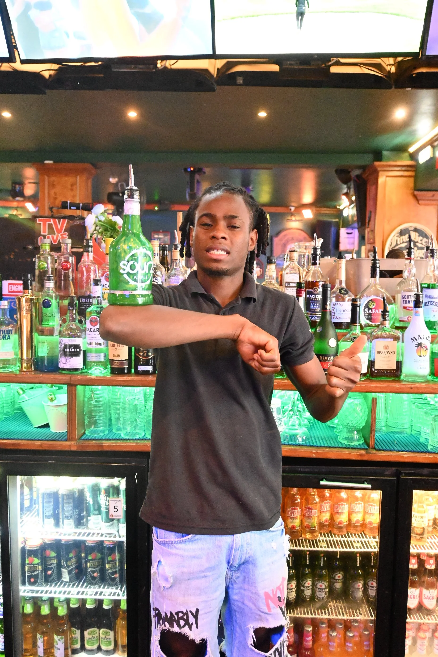 A man standing in front of a well-stocked bar or fridge, holding a green bottle with a straw, and pointing with his right hand. The background shows various bottles and cans of drinks.