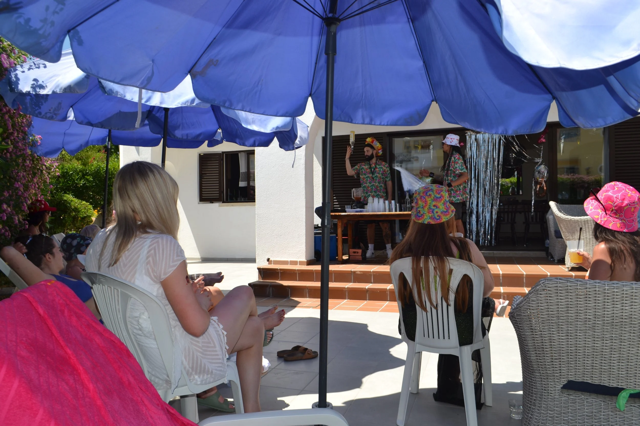 People sitting on chairs in an outdoor patio under large blue umbrellas, with a man and woman playing music behind a table. The setting appears to be a casual outdoor gathering or party.