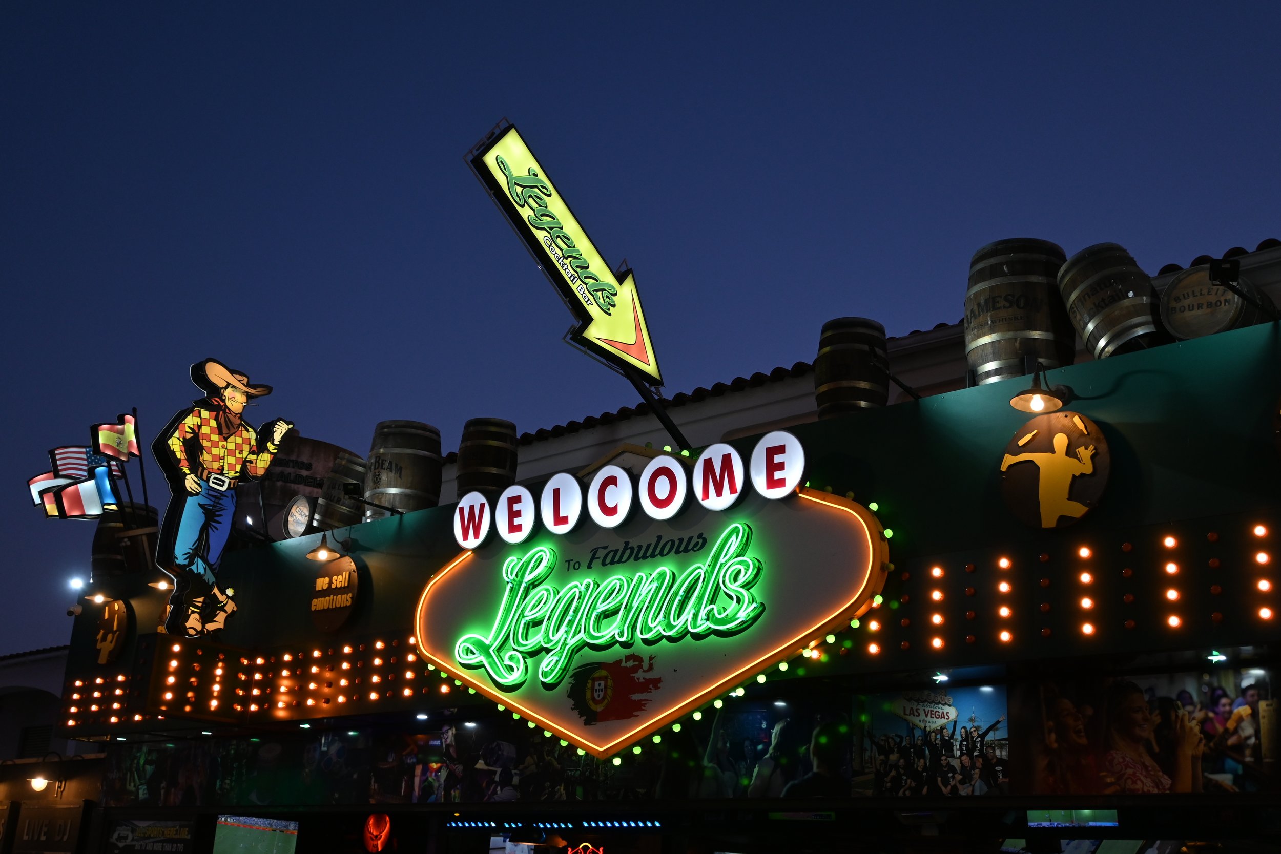 Nighttime scene of a Las Vegas casino sign that says 'Welcome to Fabulous Legends' with neon lights, a cowboy figure, waving flags, and barrels stacked on the roof.