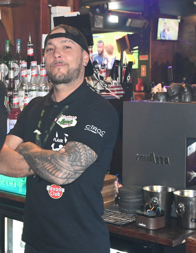 Bartender with tattoos posing in a bar, surrounded by bottles and bar equipment, with TV screens in the background