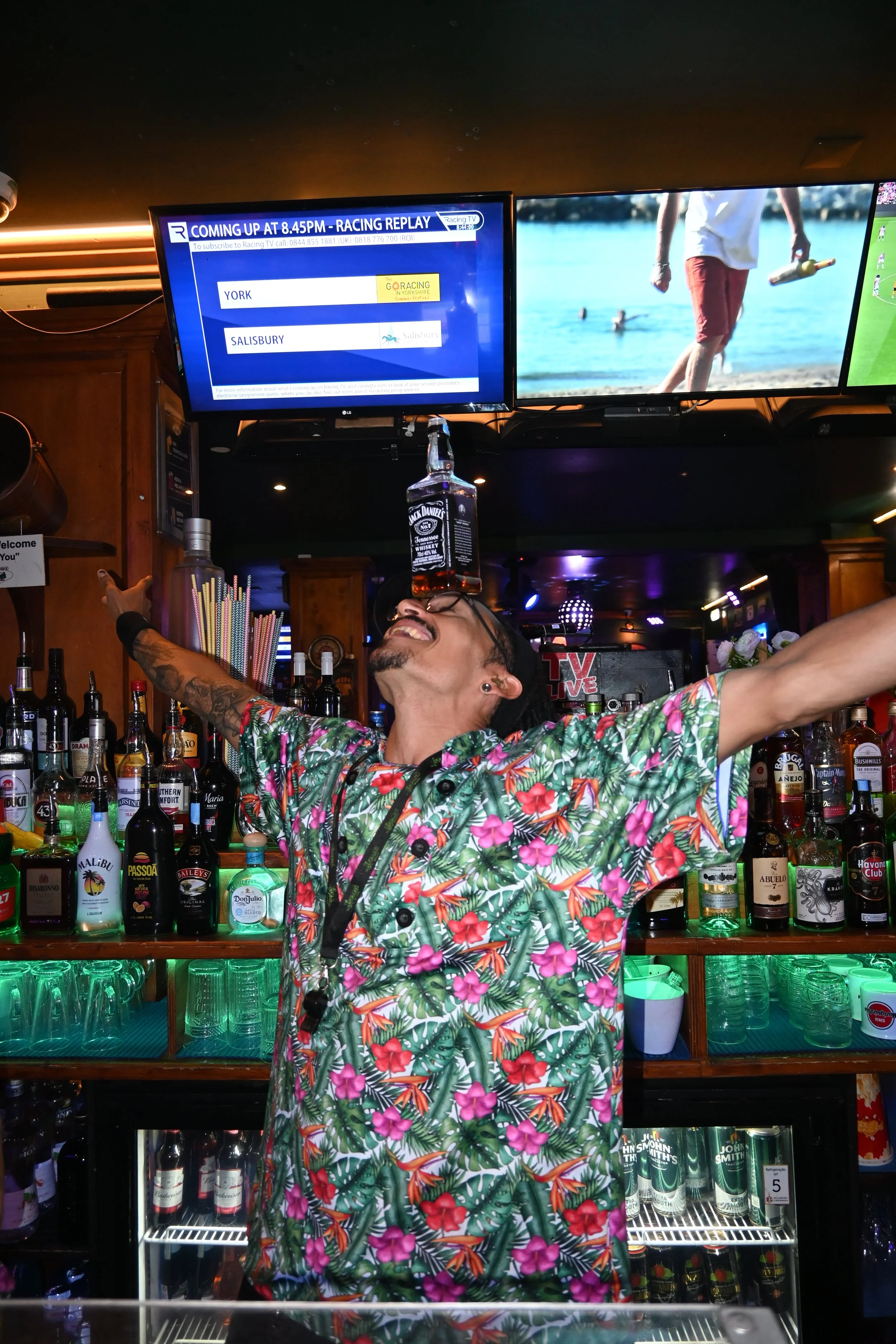 A bartender wearing a colorful tropical shirt with pink flowers and green leaves, smiling with arms outstretched behind a bar counter. Above, two television screens display a racing event and a beach scene. The bar has various bottles of alcohol and glasses, with a bottle of Jack Daniel's whiskey on top of the television.