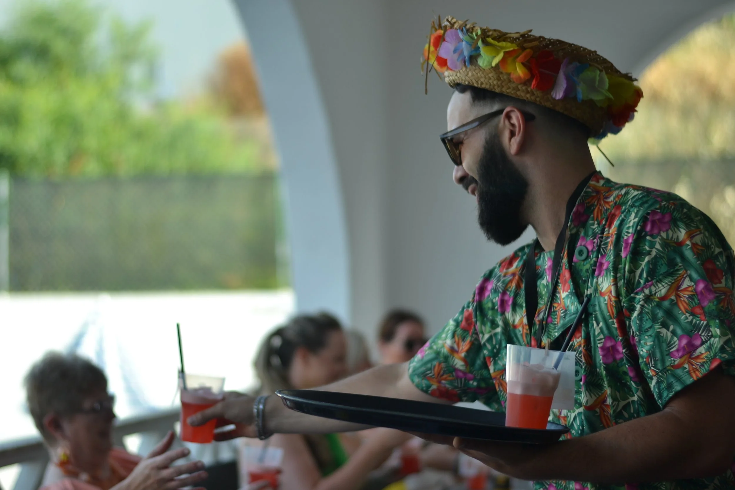 A man with a beard wearing sunglasses and a colorful Hawaiian shirt with a floral pattern is serving drinks at a party. He is wearing a large straw hat decorated with multi-colored artificial flowers.