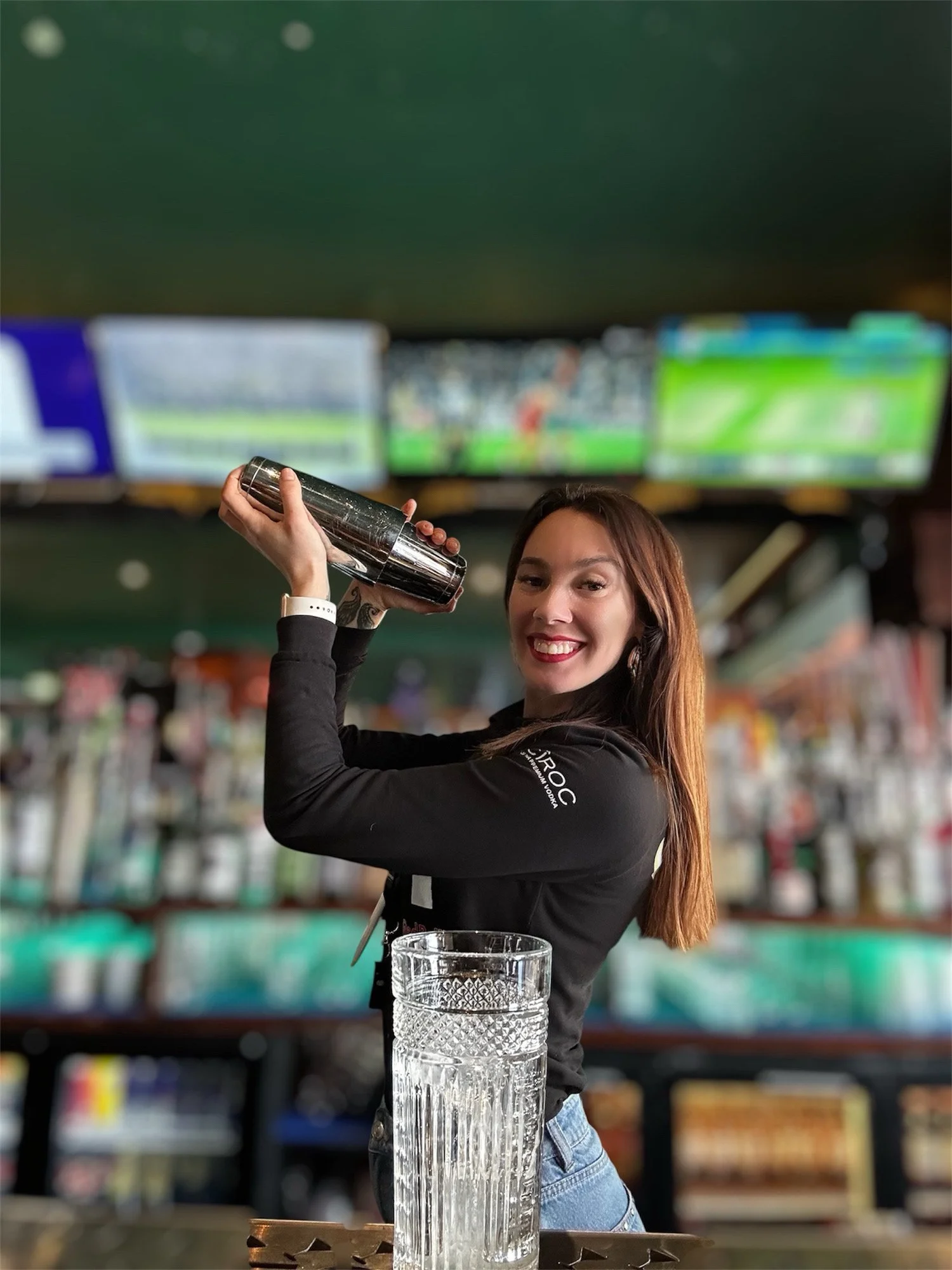 A woman with long brown hair smiling and holding a cocktail shaker over a glass in a bar or pub. Blurred televisions or screens showing sports events in the background.