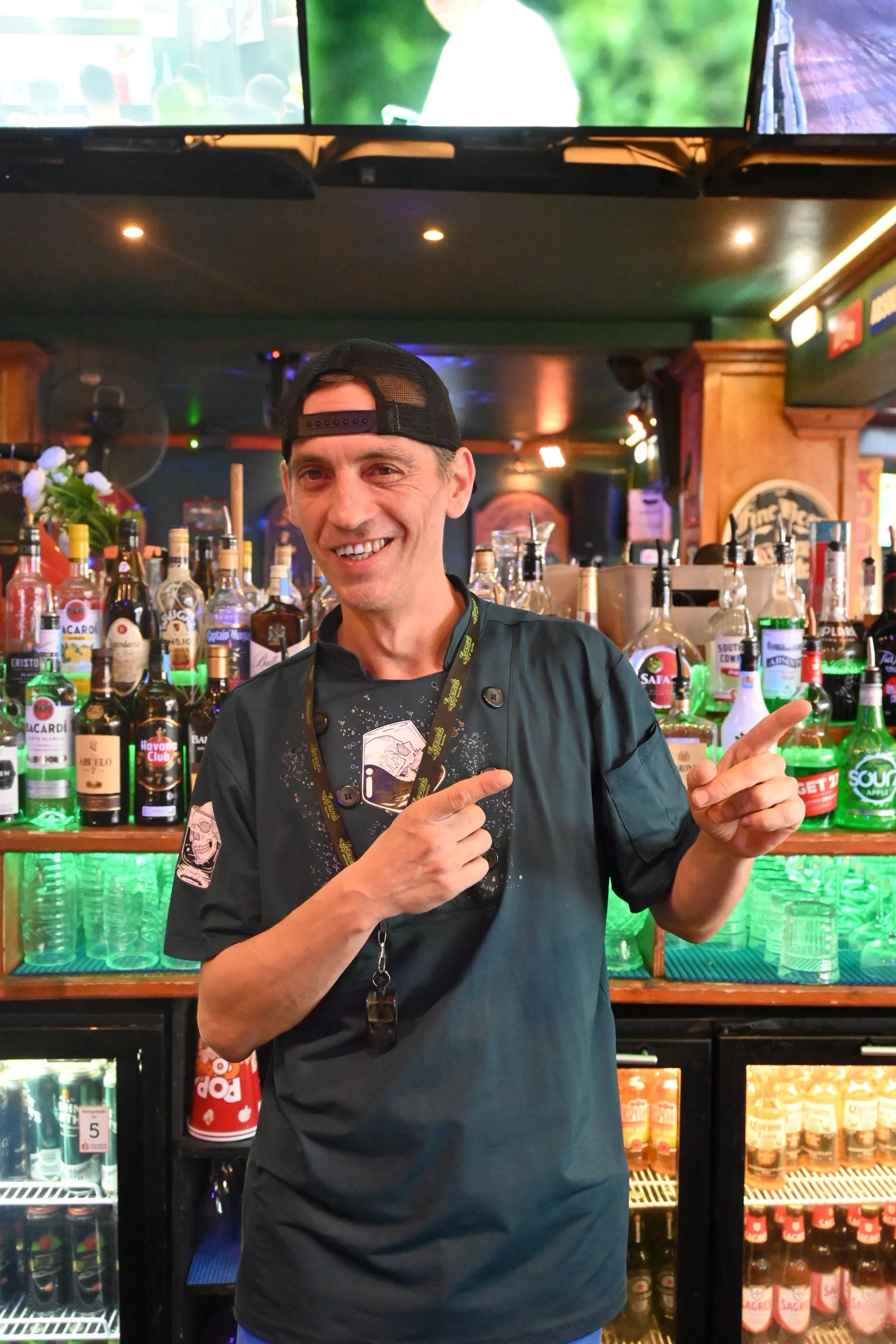 A smiling man in a black chef's jacket and a backwards black cap standing in front of a bar with various bottles of alcohol and a fridge behind him, in a pub or bar setting.
