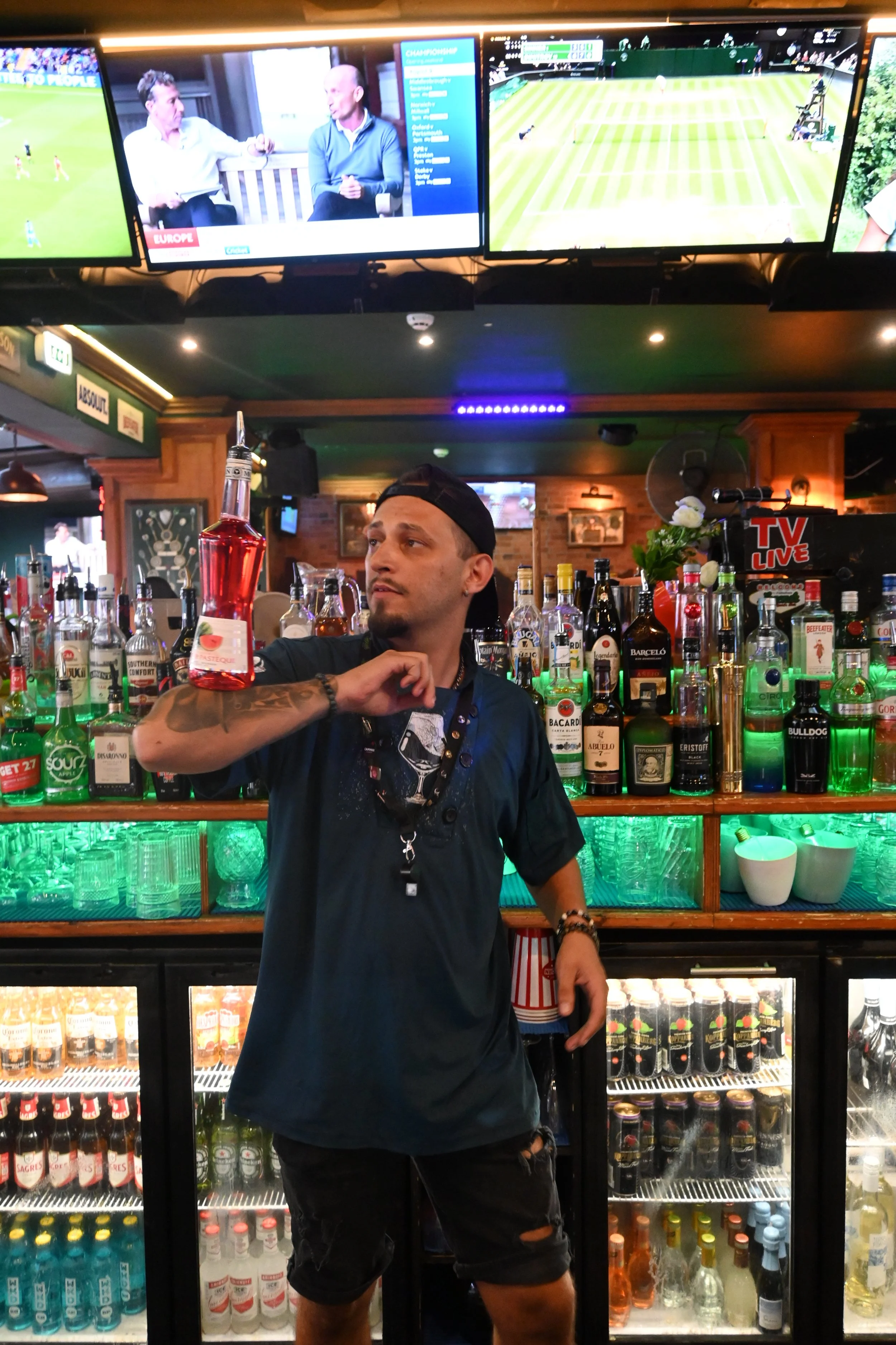 A bartender in a bar holding a red liquor bottle, surrounded by various bottles of alcohol and glassware, with multiple TV screens displaying sports and news in the background.