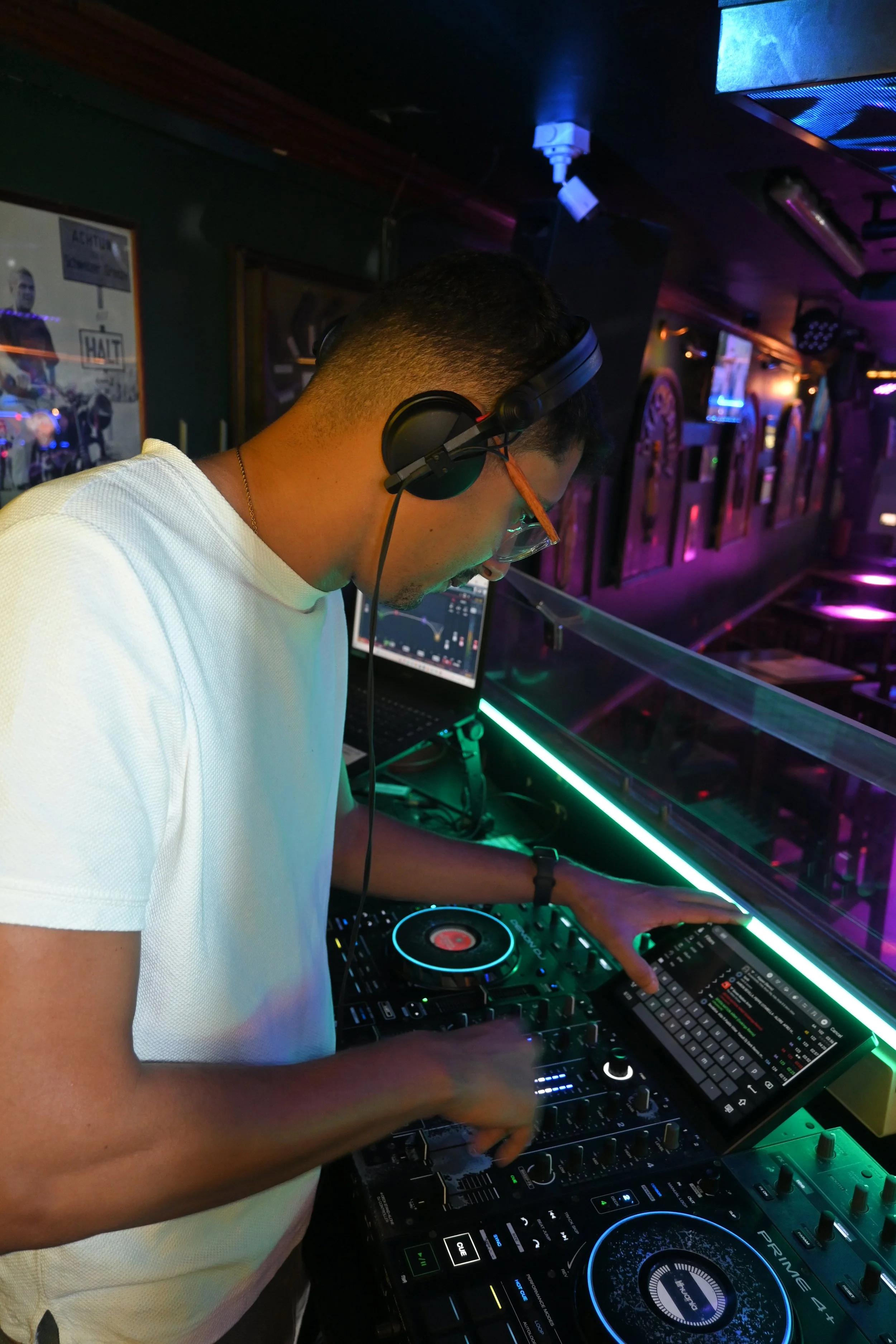 A DJ wearing glasses and headphones spinning music at a nightclub with colorful lighting and a DJ controller.