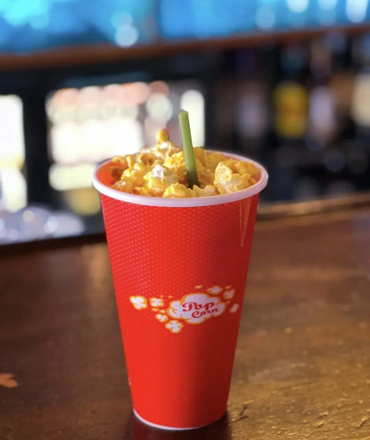 A red paper cup filled with popcorn and a green straw on a wooden table.