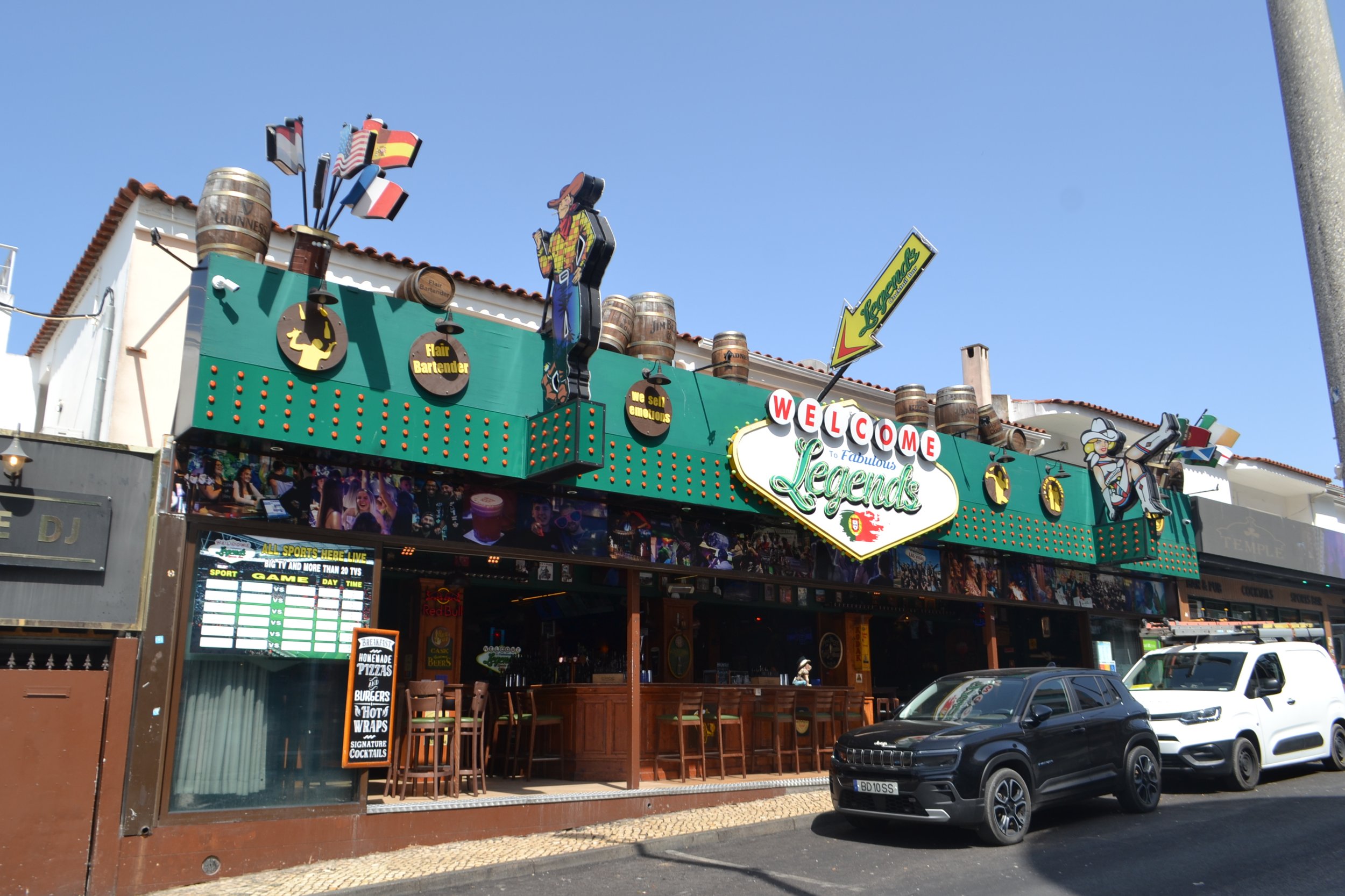 Exterior of a bar or restaurant with a colorful sign that says 'Welcome to Fabulous Legends' and various cartoon signs on the facade, including a cowboy, a cowgirl, and a waitress, with cars parked in front.