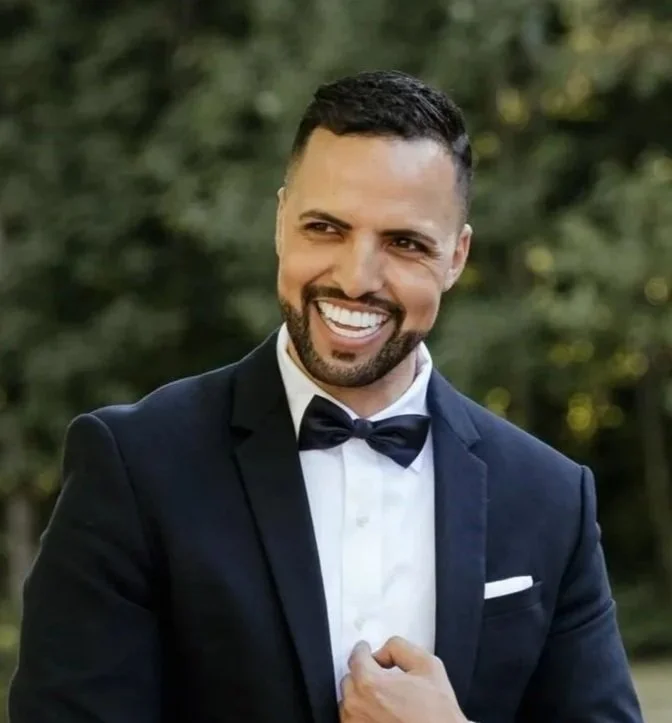 A smiling man in a tuxedo with a bow tie, outdoors with blurred greenery in the background.