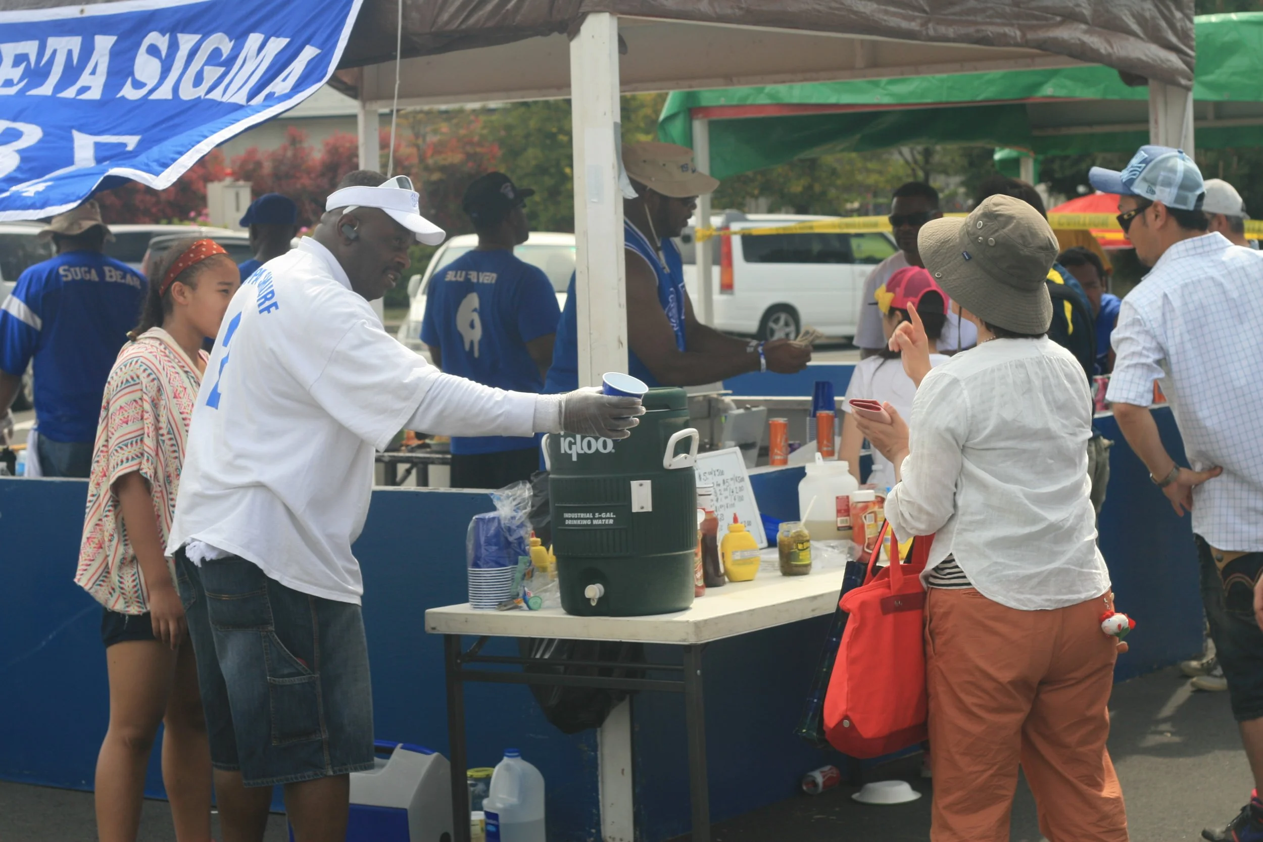 People lined up at a food stand outdoors, some holding cups, with tents and vehicles in the background.