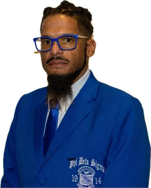 A man with a beard and glasses wearing a blue blazer with the Pi Beta Sigma fraternity emblem embroidered on it.