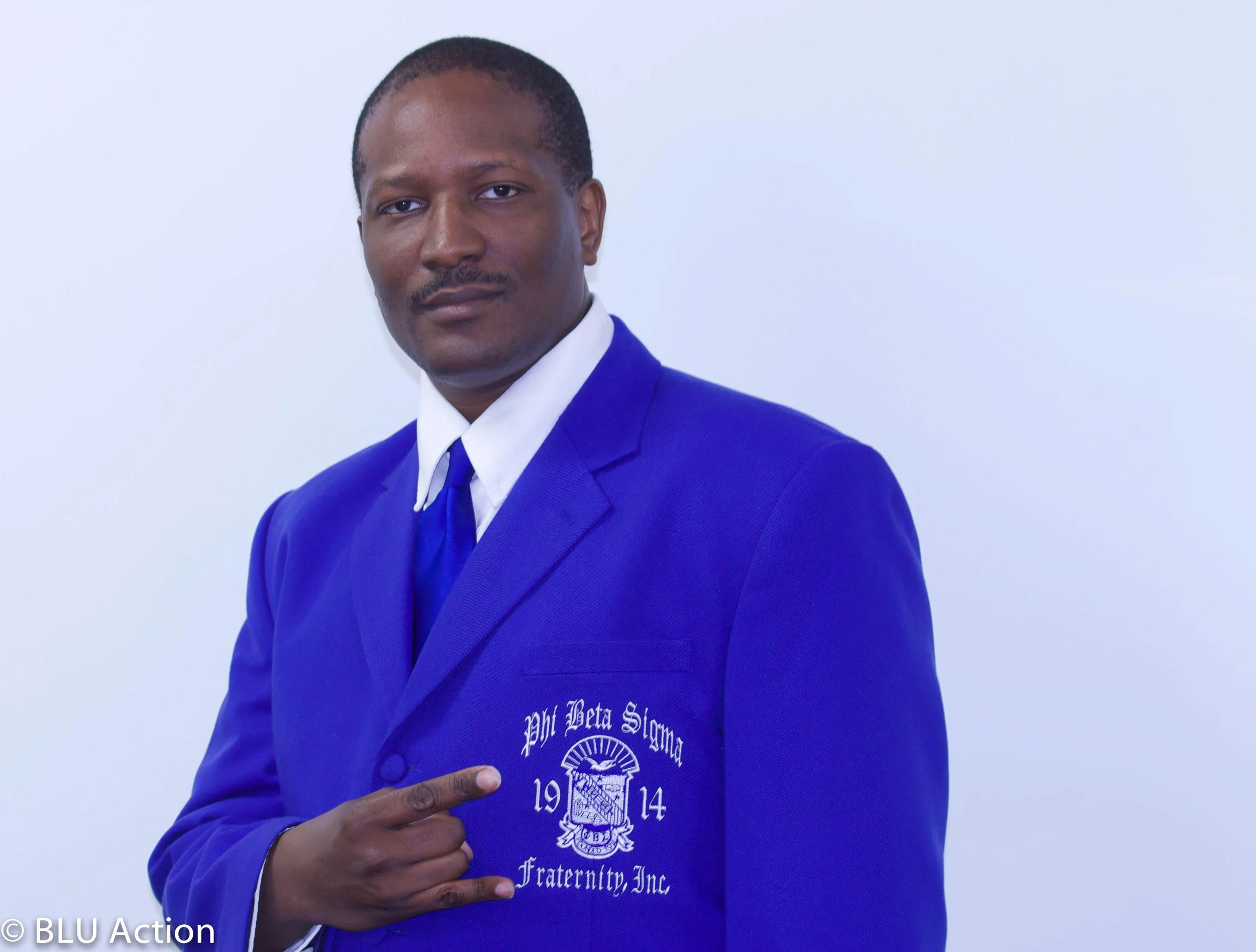 A man in a blue blazer pointing to a logo on his blazer, with a serious expression, against a plain white background.