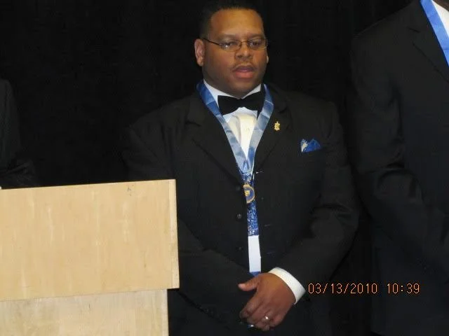 A man dressed in a black tuxedo with a bow tie, wearing medals and a blue ribbon, standing at a podium during a formal event.