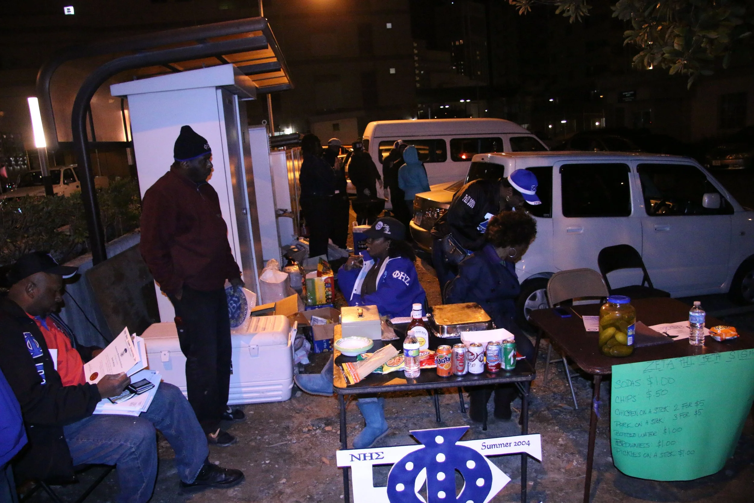 People gathered at a small outdoor food stand at night, with tables displaying canned drinks, snacks, and food. Some are sitting and reading, while others are waiting or working at the stand, which has a sign with prices for sodas, chips, chicken on a stick, and pickles.