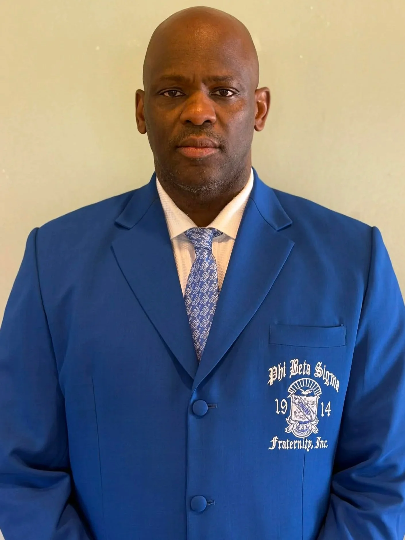 A bald man dressed in a blue blazer with patch on the chest, white shirt, and patterned tie, standing against a plain light background.