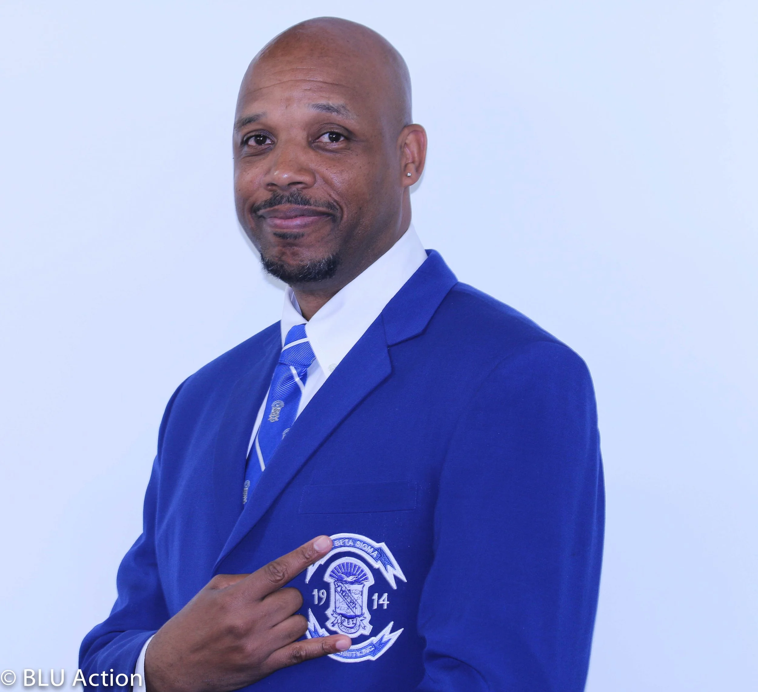 A man in a blue suit and tie pointing at a patch on his blazer, standing against a plain white background.