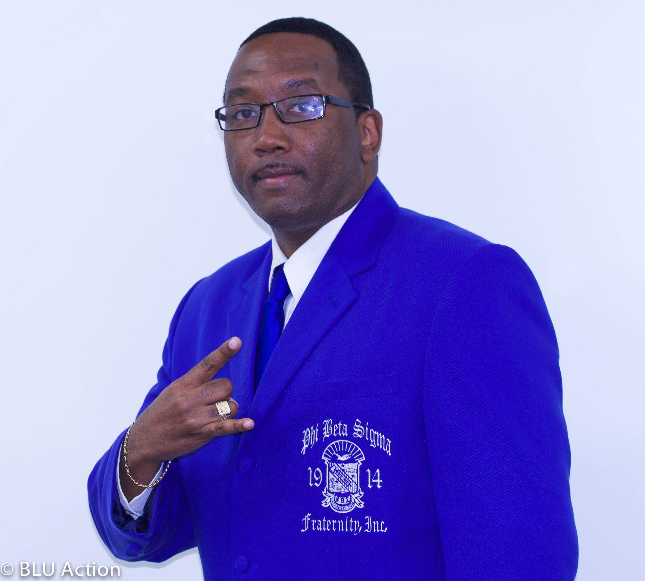 A man wearing a blue blazer with white embroidery, glasses, and a blue tie, pointing to the blazer with his right hand, standing against a plain white background.