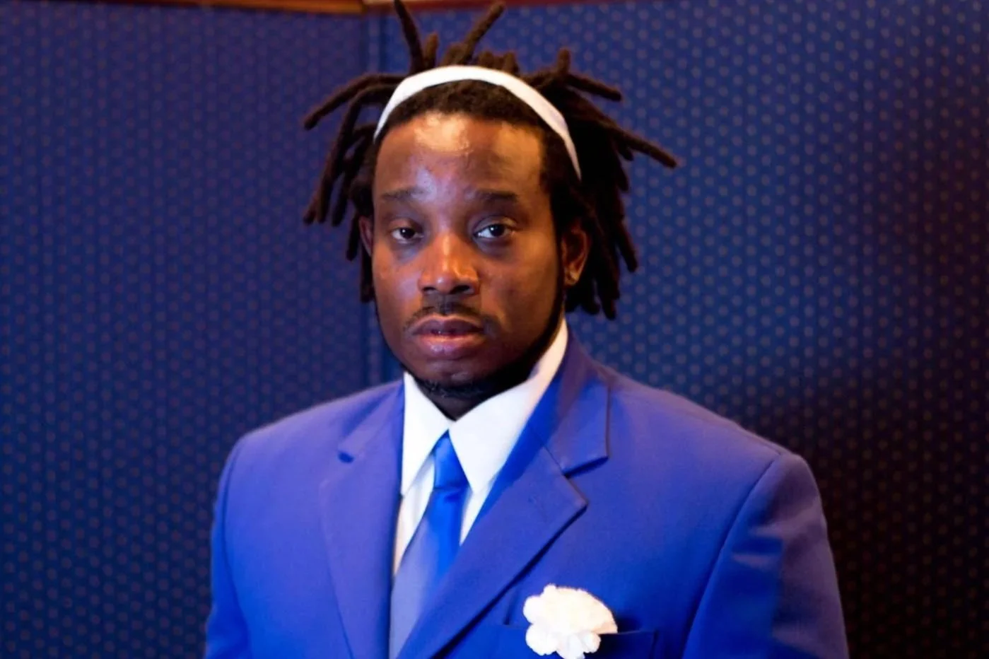 Man with dreadlocks wearing a blue suit and white shirt, with a white flower in his pocket, standing against a blue patterned background.