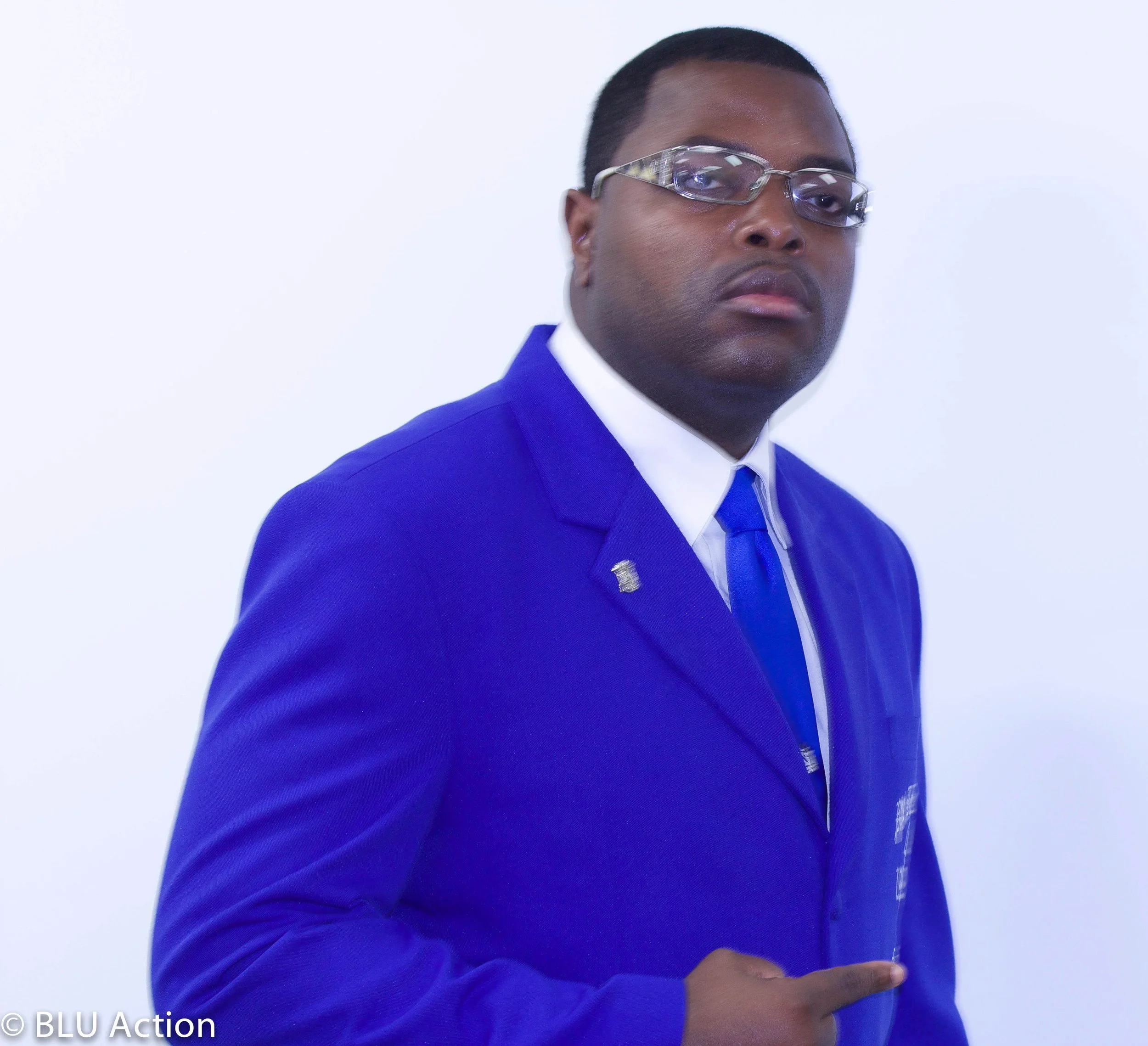 A man in a blue suit and glasses pointing to a badge on his chest against a white background.