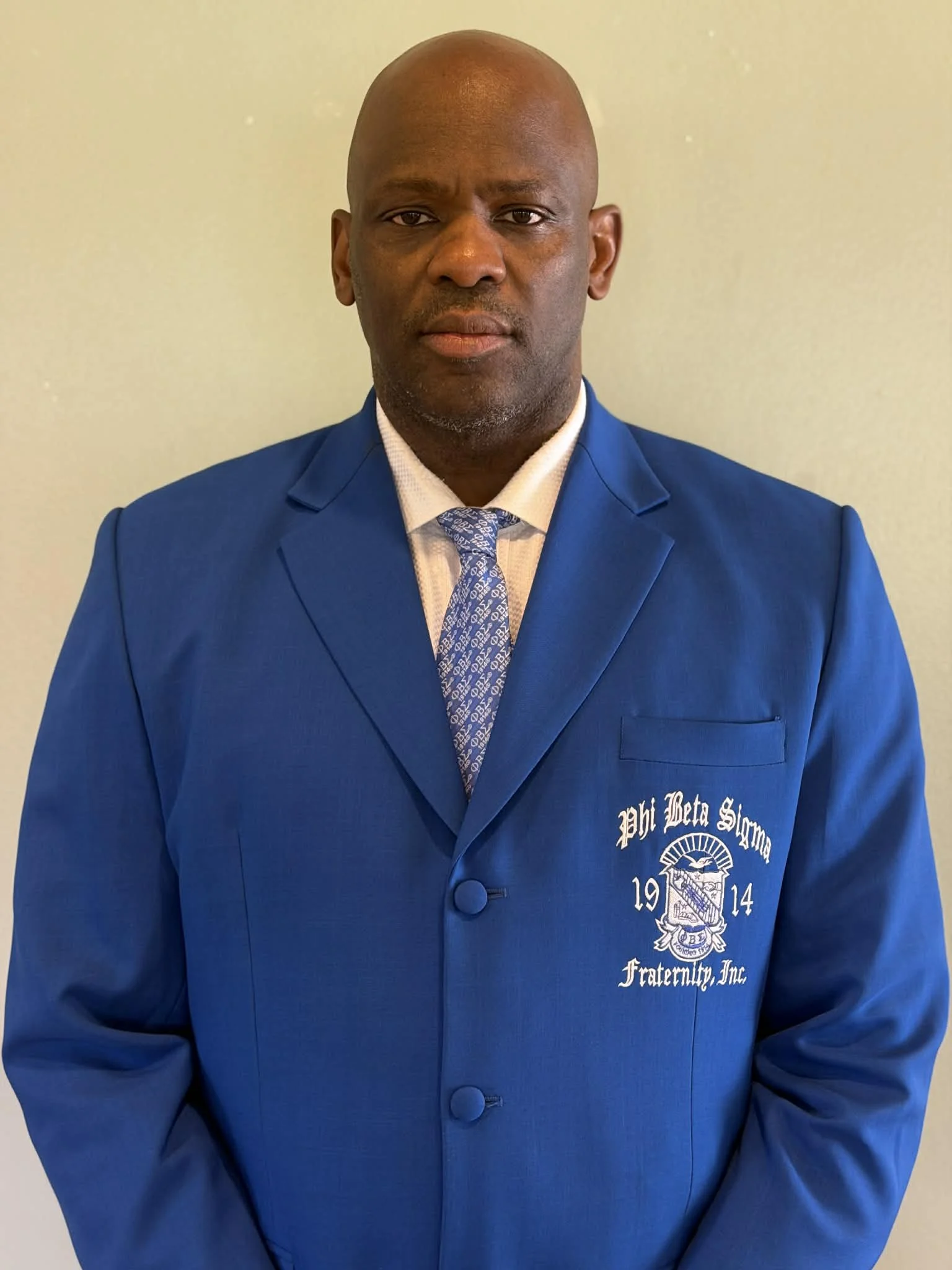 A man in a blue blazer with pi beta sigma fraternity insignia on the chest, white shirt, and patterned tie, standing against a plain background.