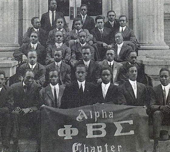 Group photo of young African American men in suits holding a banner with American Greek fraternity symbols and text on a set of steps.