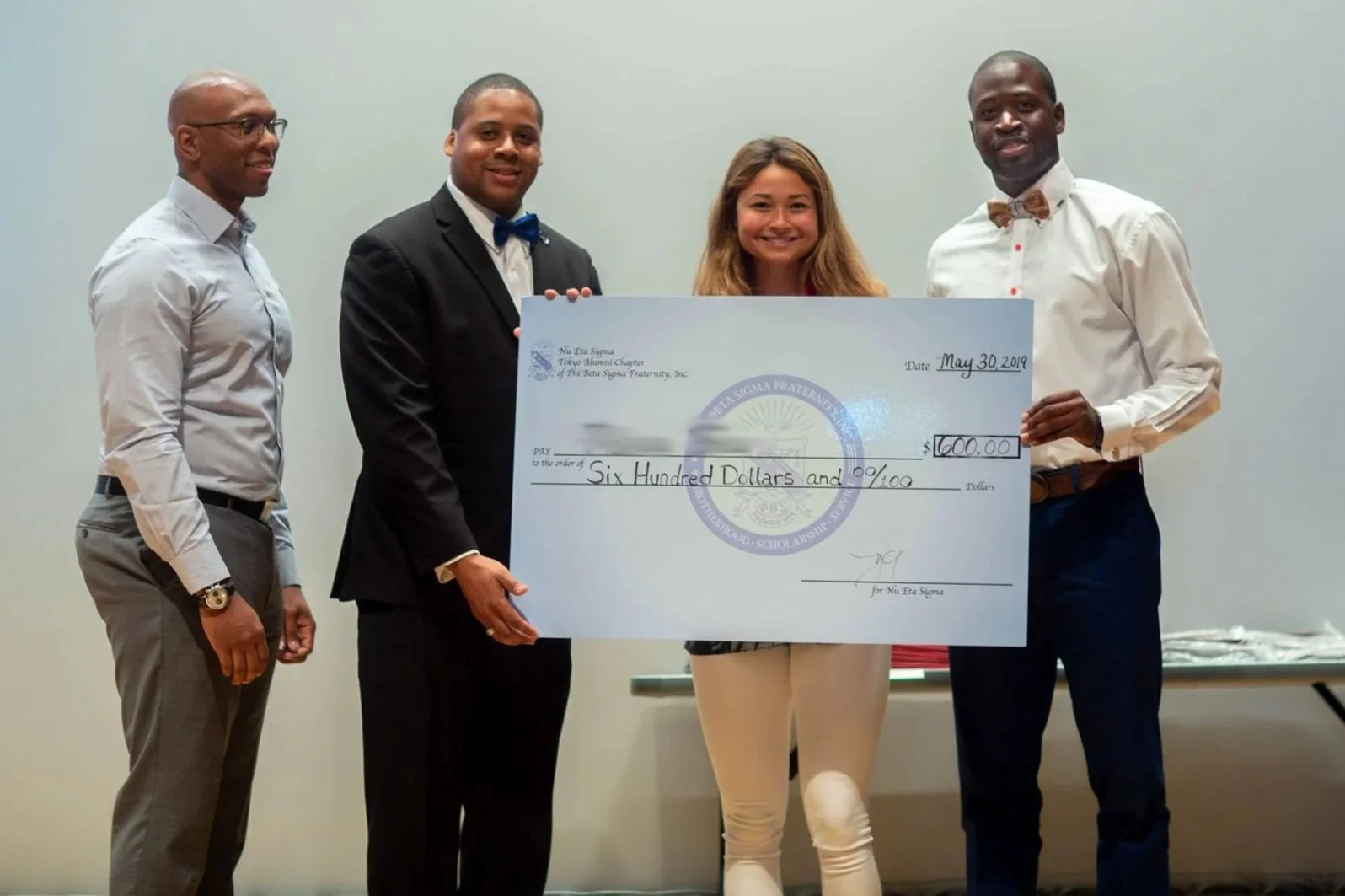 Four individuals standing together holding an oversized check for $600, awarded by Pi Eta Sigma Fraternity on May 30, 2019. The group includes two men in suit and dress shirt, one woman with long hair, and another man wearing a bow tie and white shirt. They are inside a room with a plain wall background.