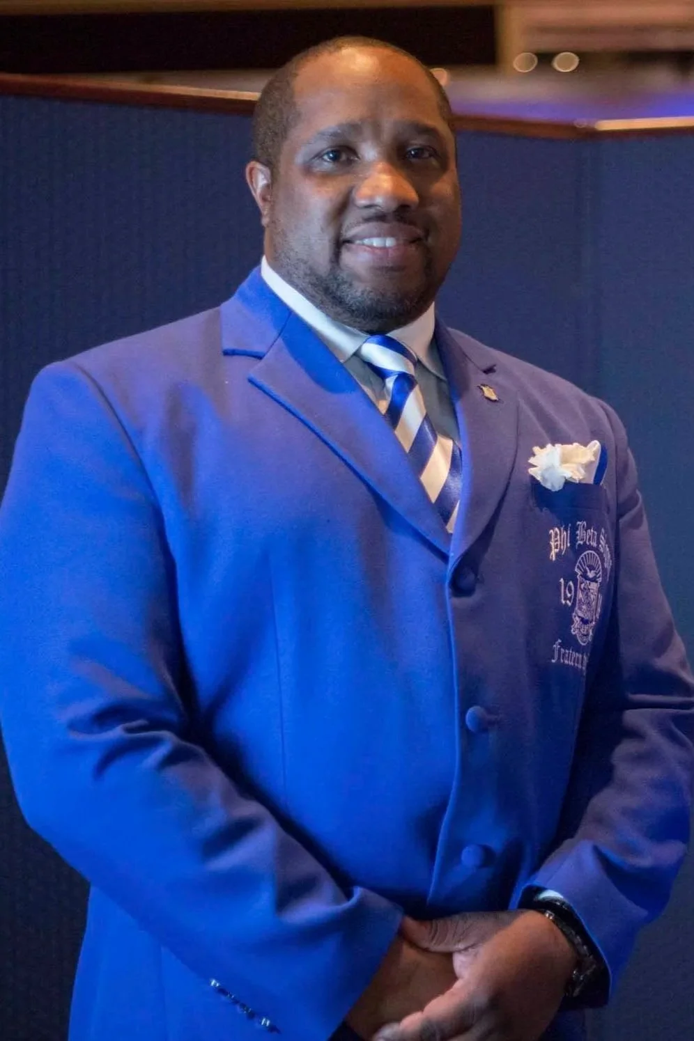 Man in a blue suit with a matching striped tie and a white pocket square, standing in front of a dark blue background, smiling at the camera.