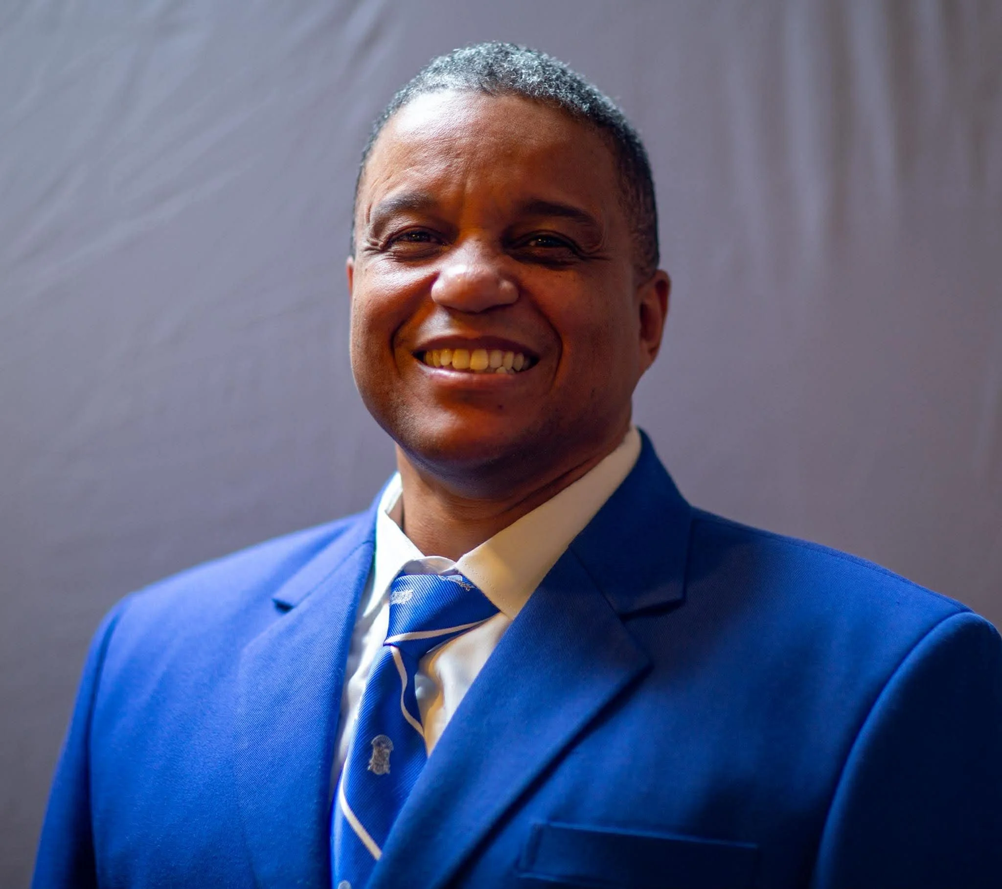A smiling man in a blue suit and tie against a plain background.