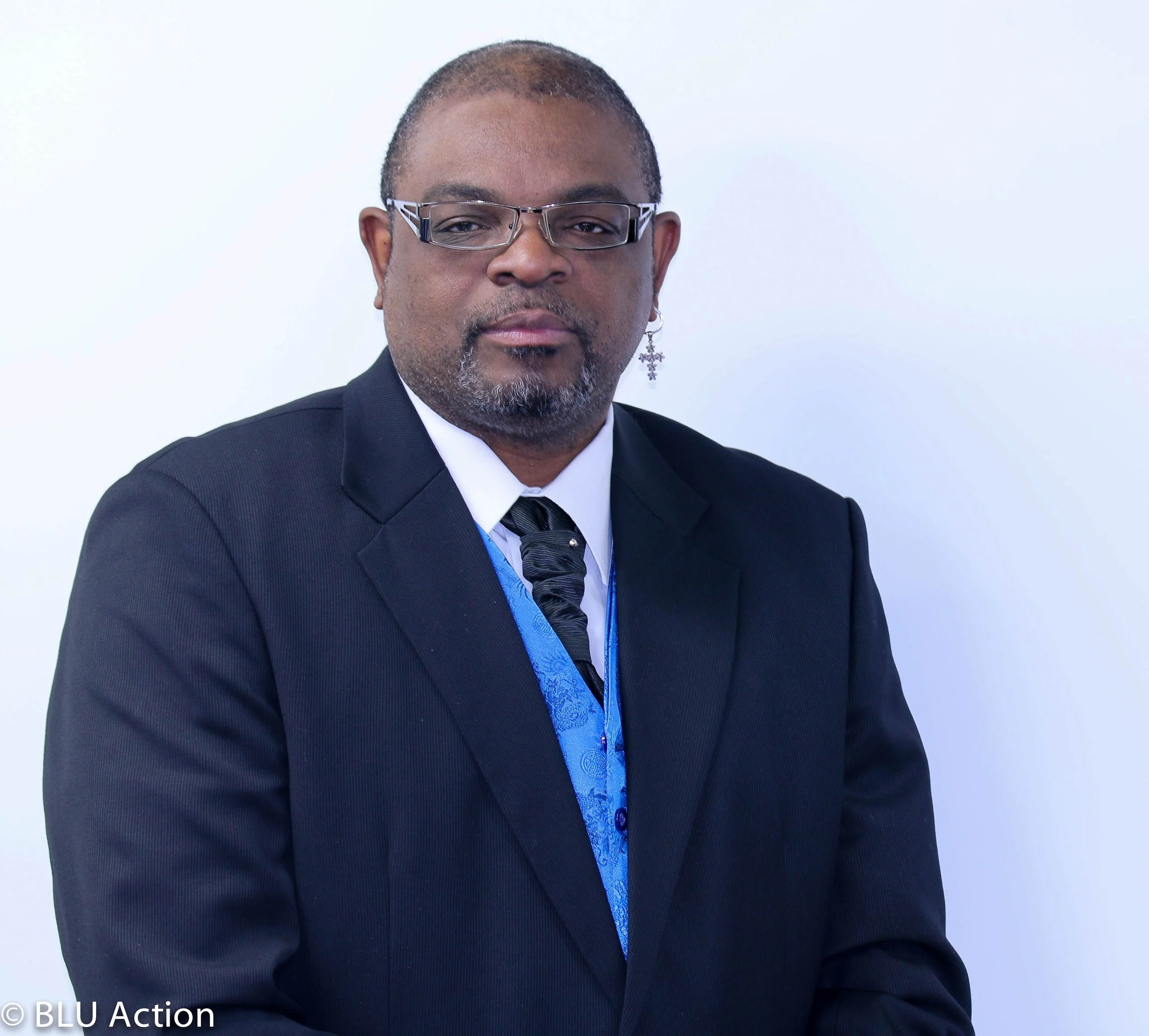 A man with glasses, earring, and a goatee, wearing a black suit and tie, standing against a white background.