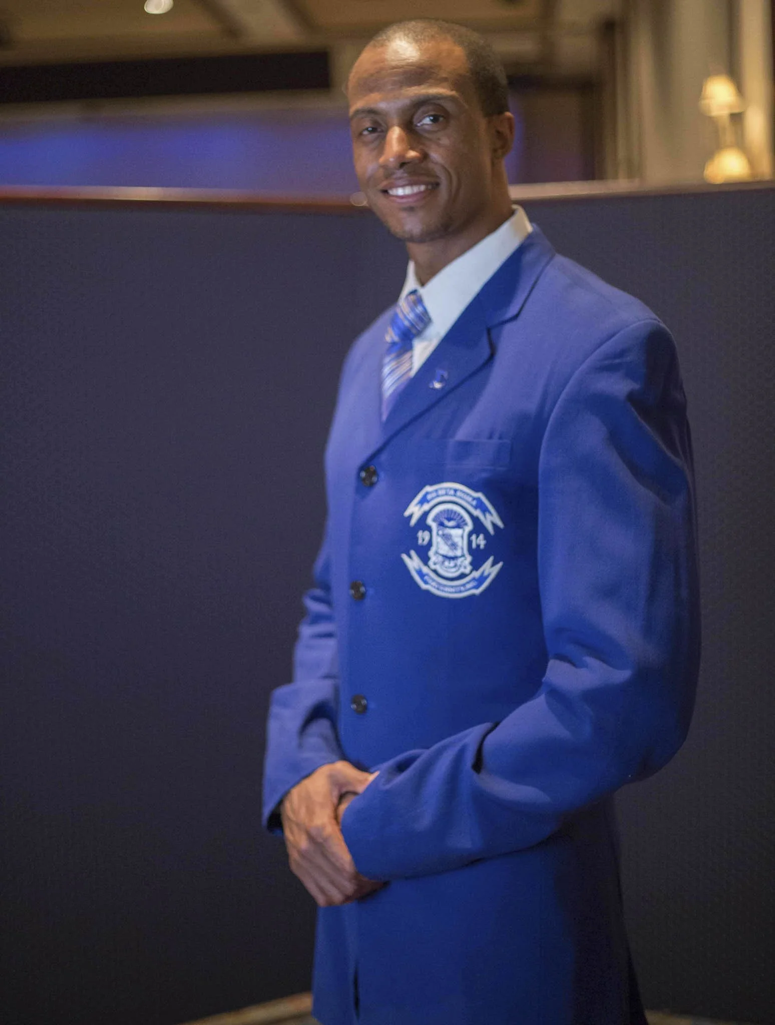 A man in a blue blazer with an emblem, white shirt, and striped tie standing indoors, smiling at the camera.