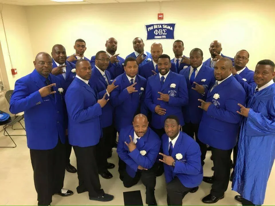 A group of men dressed in blue suits with white flowers on the lapels posing indoors. Some are making hand gestures, and there is a Phi Beta Sigma fraternity banner on the wall.