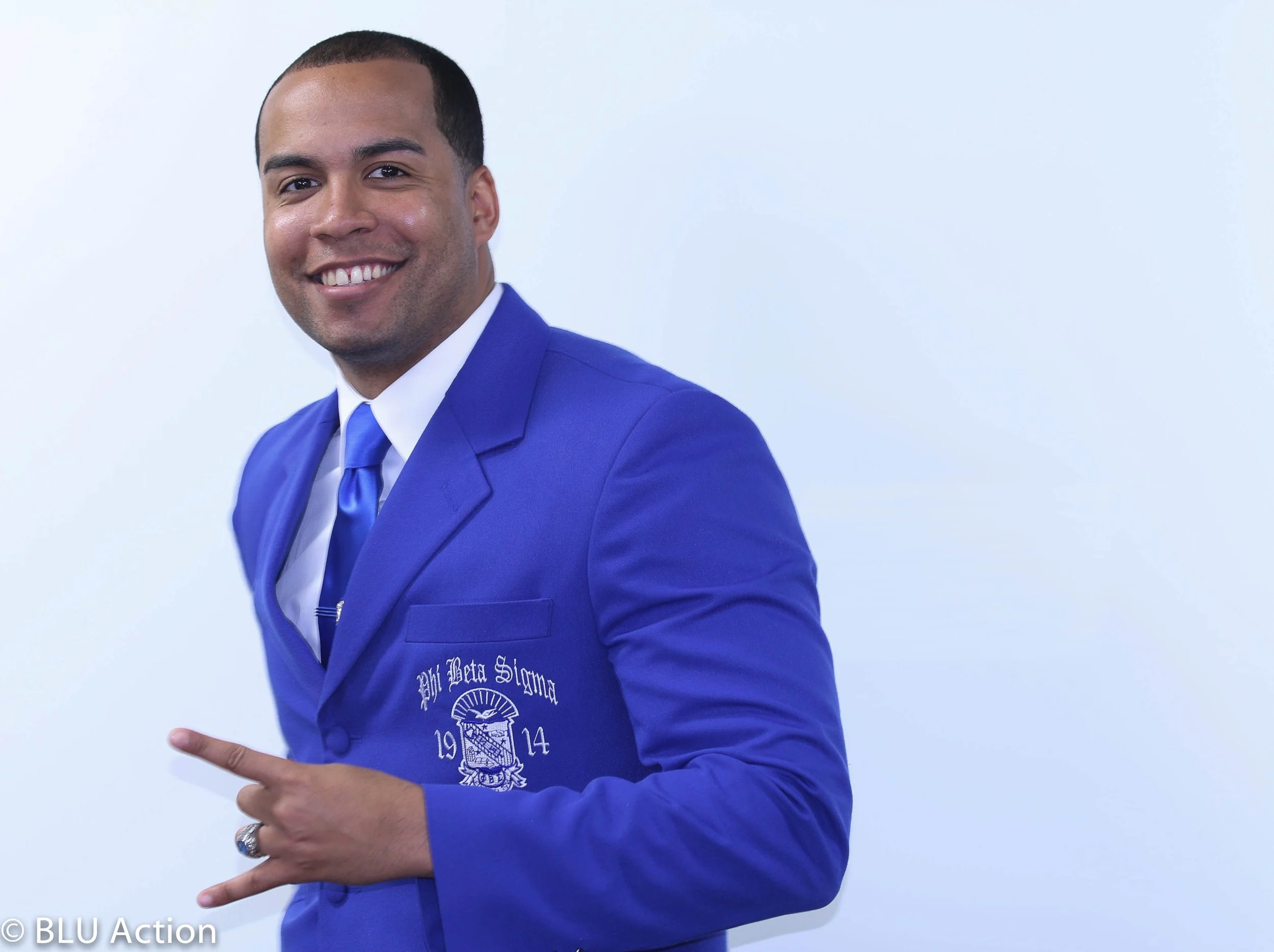 A smiling man wearing a blue blazer, white dress shirt, and blue tie pointing to his blazer with his right hand, standing against a plain light background.
